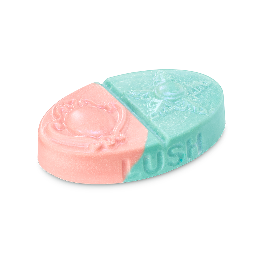 Oval body balm split diagonally into pink and teal sections with embossed brooch and star designs. Features the 'LUSH' logo on the side with a fine shimmer.