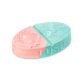 Oval body balm split diagonally into pink and teal sections with embossed brooch and star designs. Features the 'LUSH' logo on the side with a fine shimmer.