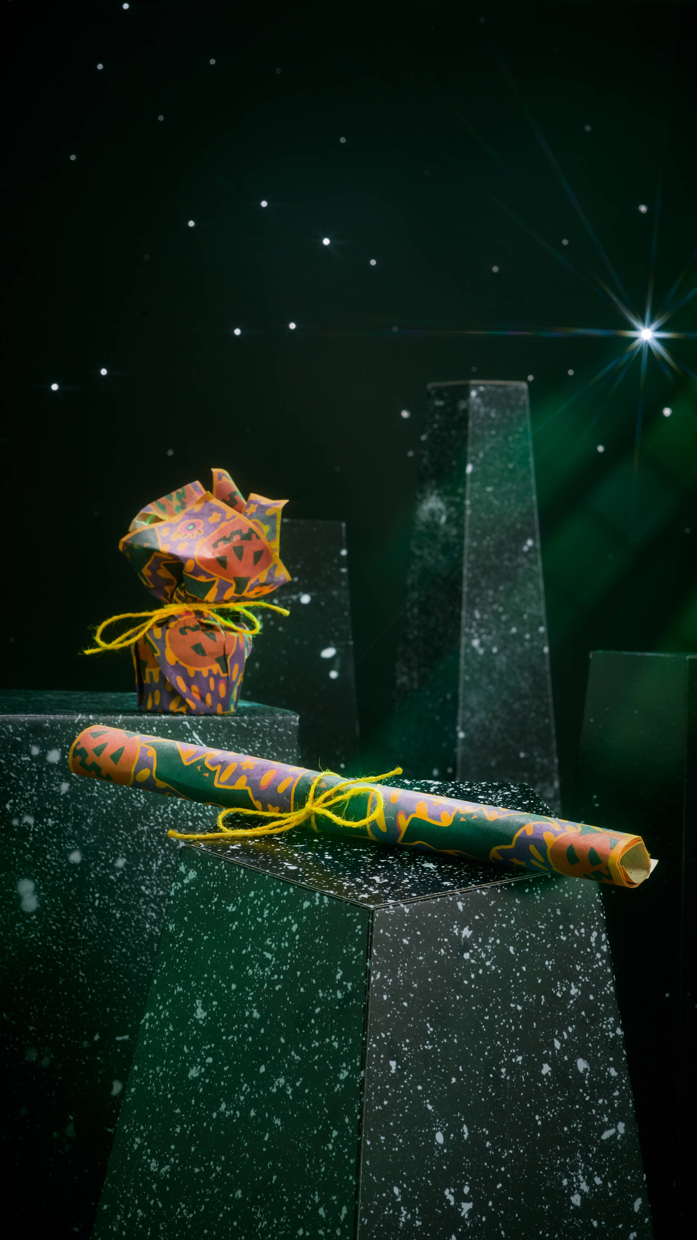 A rolled sheet of patterned Lokta paper and a small gift wrapped in patterned Halloween paper and tied with yellow string, rest on dark, speckled pillars. The background is a dark starry night sky with a bright, shining star.