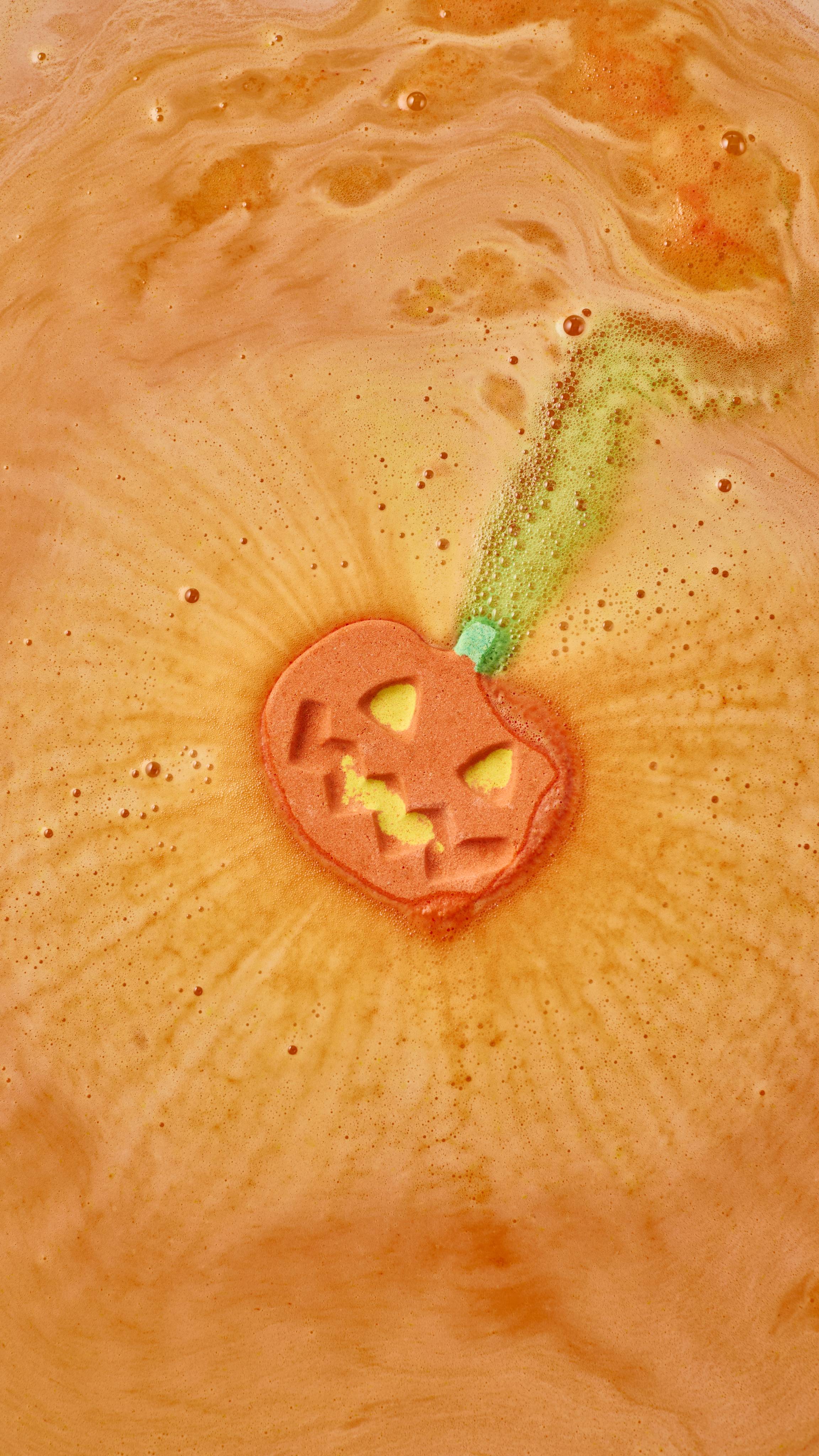 An orange pumpkin-shaped bath bomb dissolves in a bath, shooting a trail of bubbly green foam from its stalk. Concentric ripples of fine orange froth radiate outward from the carved jack-o'-lantern face into the warm orange water.