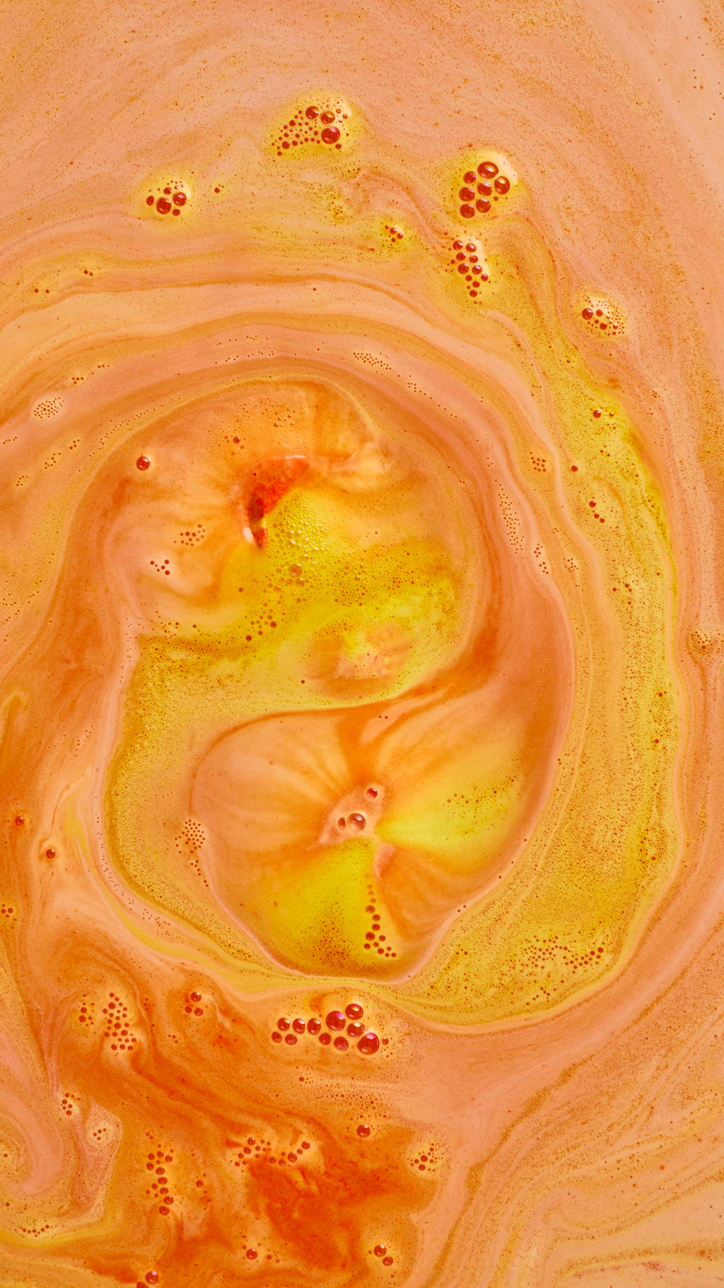 An abstract aerial shot of bath bomb foam swirling in water, creating a large spiral pattern of bright yellow and warm, creamy orange froth. Clusters of fine, orange-tinted soapy bubbles are scattered across the textured surface.