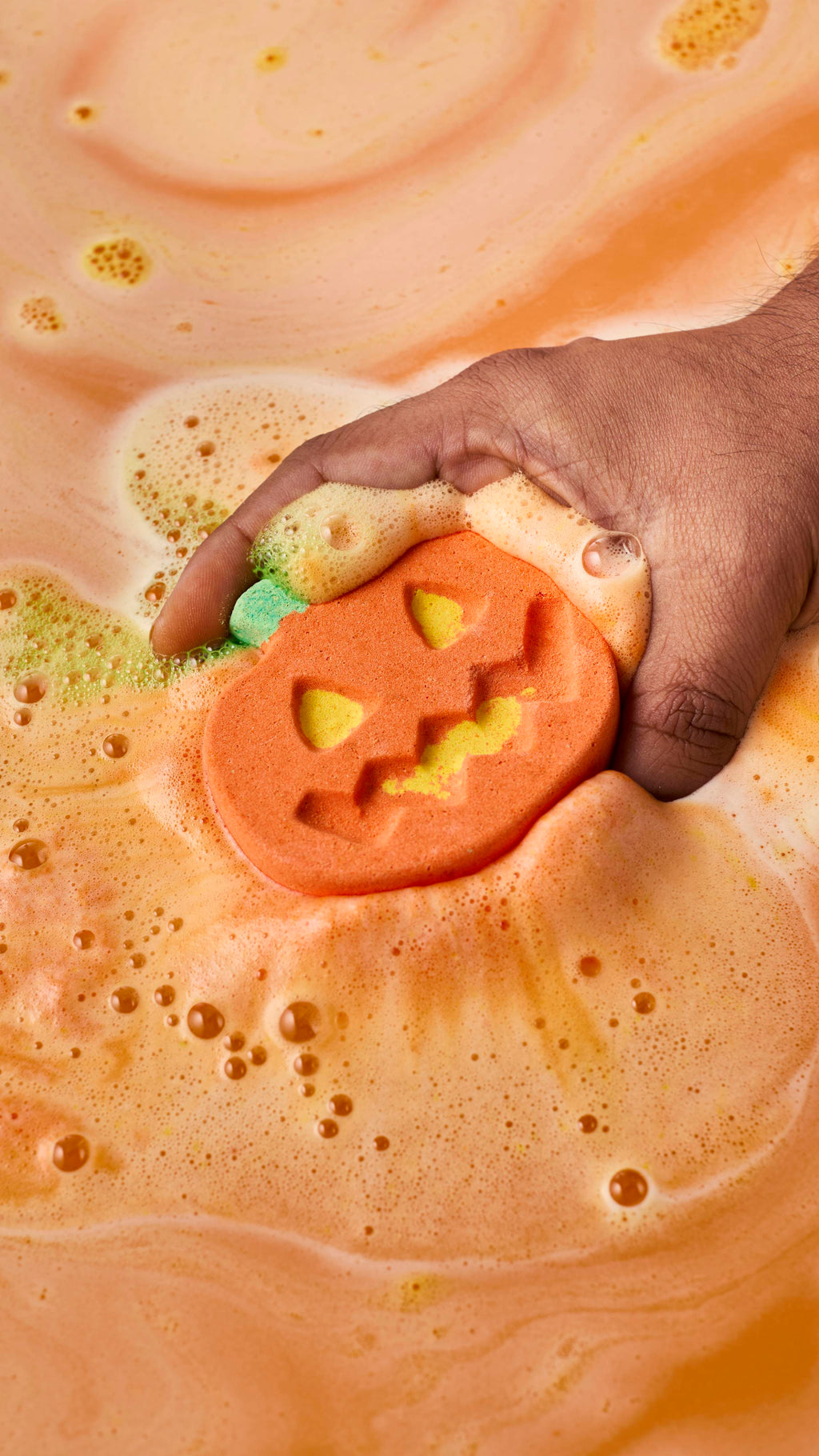 A hand places an orange pumpkin-shaped bath bomb into the water, where it begins to fizz. Creamy orange, yellow, and lime-green foam swells around the carved jack-o'-lantern face as the bath water turns a warm, opaque orange.