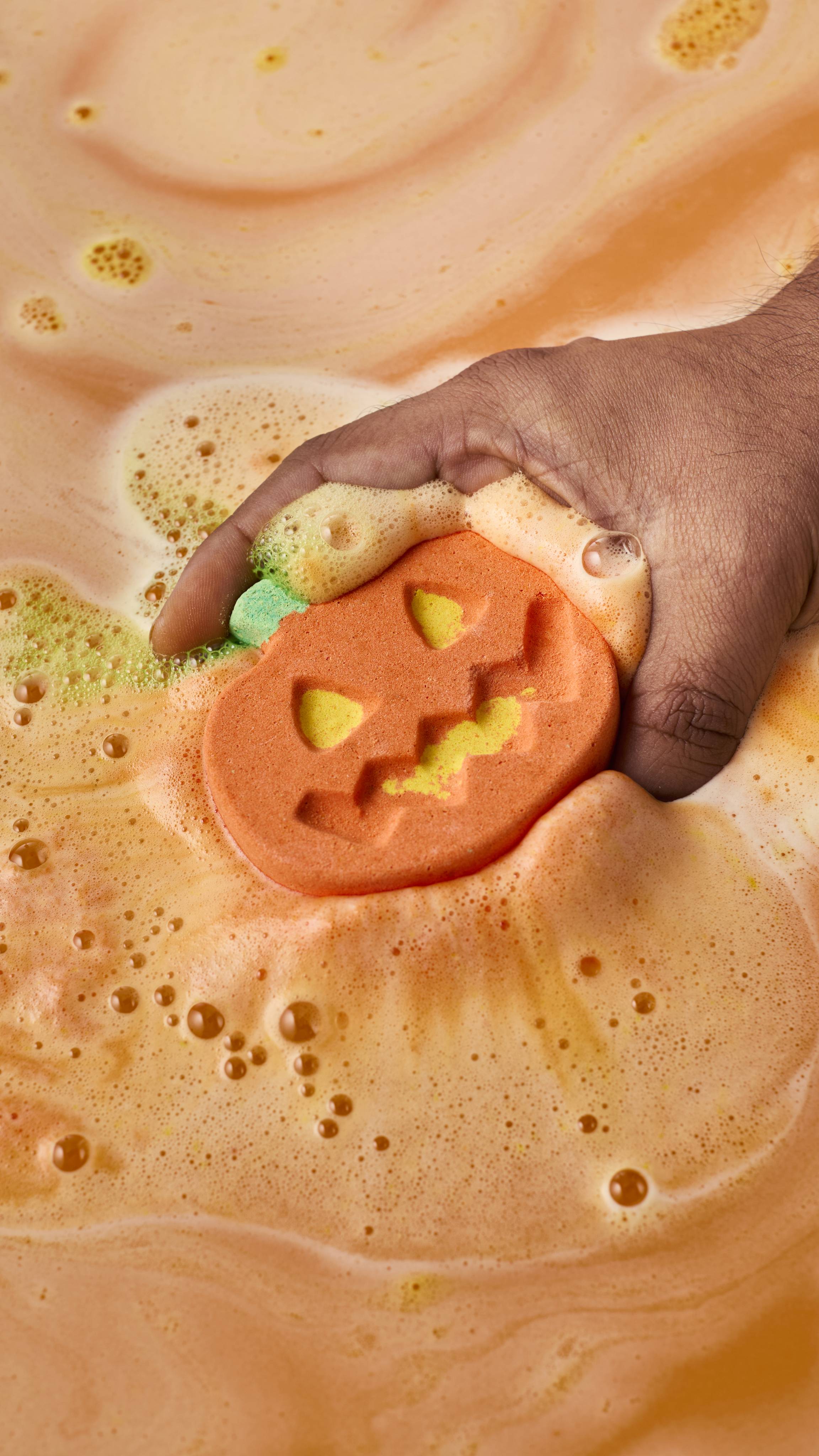 A hand places an orange pumpkin-shaped bath bomb into the water, where it begins to fizz. Creamy orange, yellow, and lime-green foam swells around the carved jack-o'-lantern face as the bath water turns a warm, opaque orange.