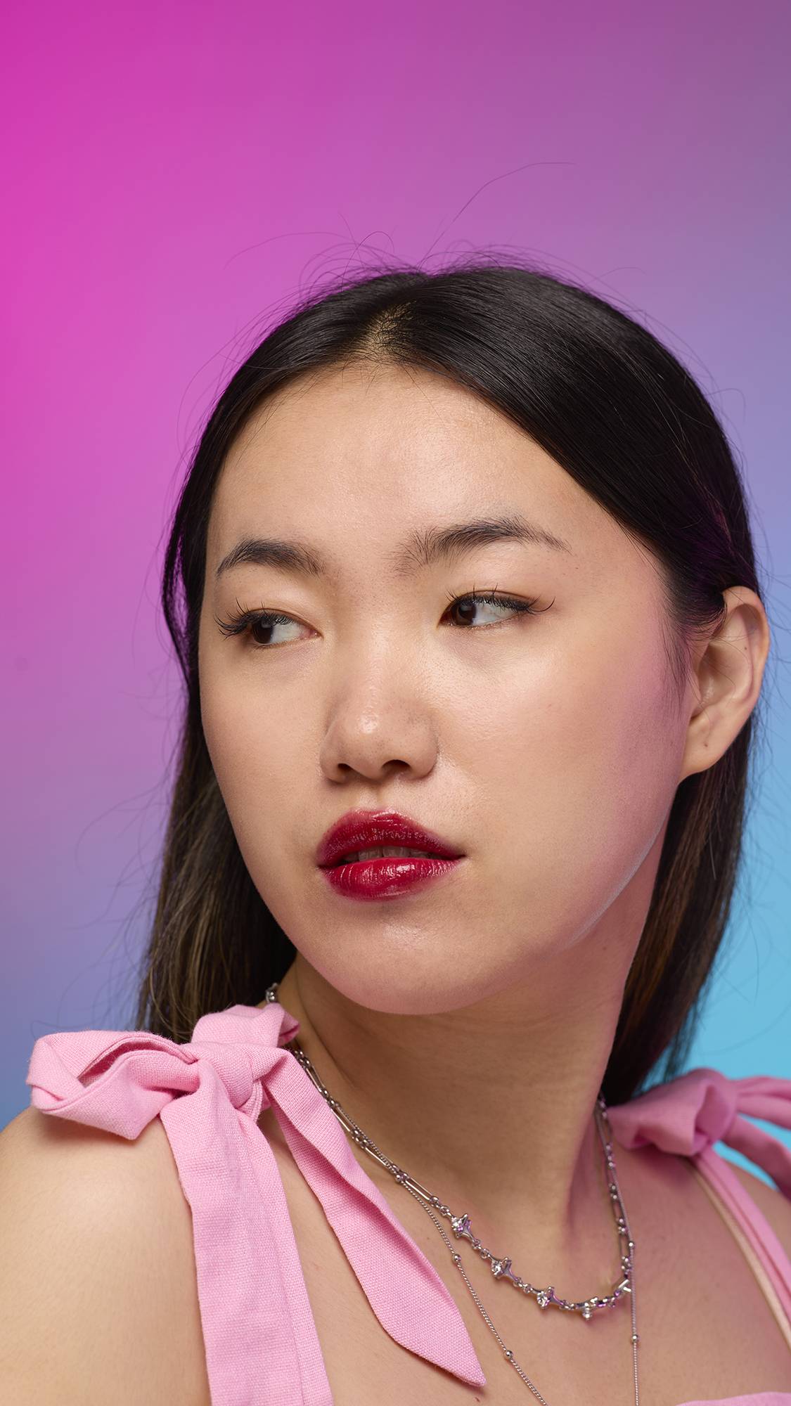 The model has long, straight dark hair and is on a pastel pink, blue and purple hue background. Their make-up has been done with the focal point being the juicy coating of carmine-red tint on their lips created by the Red Maple lip jelly.