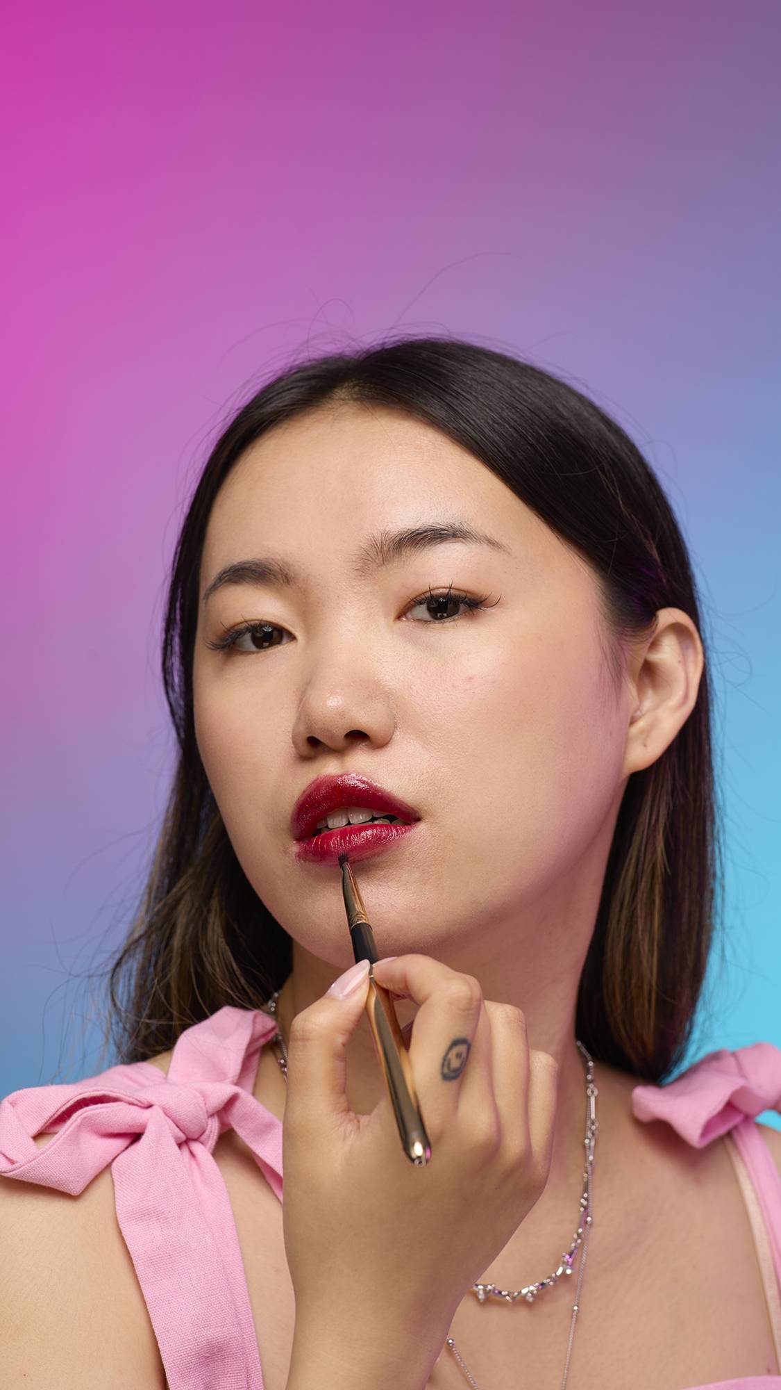 The model has long, dark hair and is on a pastel pink, blue and purple hue background. They are holding a thin, long brush to their face as they apply the vivid, Red Maple lip jelly.