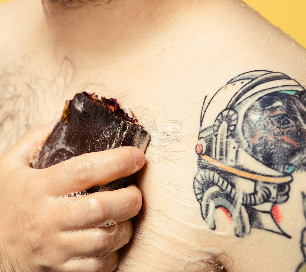 A person with tattoos sweeps Ro's Argan, a deep purple soap, topped with red and yellow rose petals, across their chest.