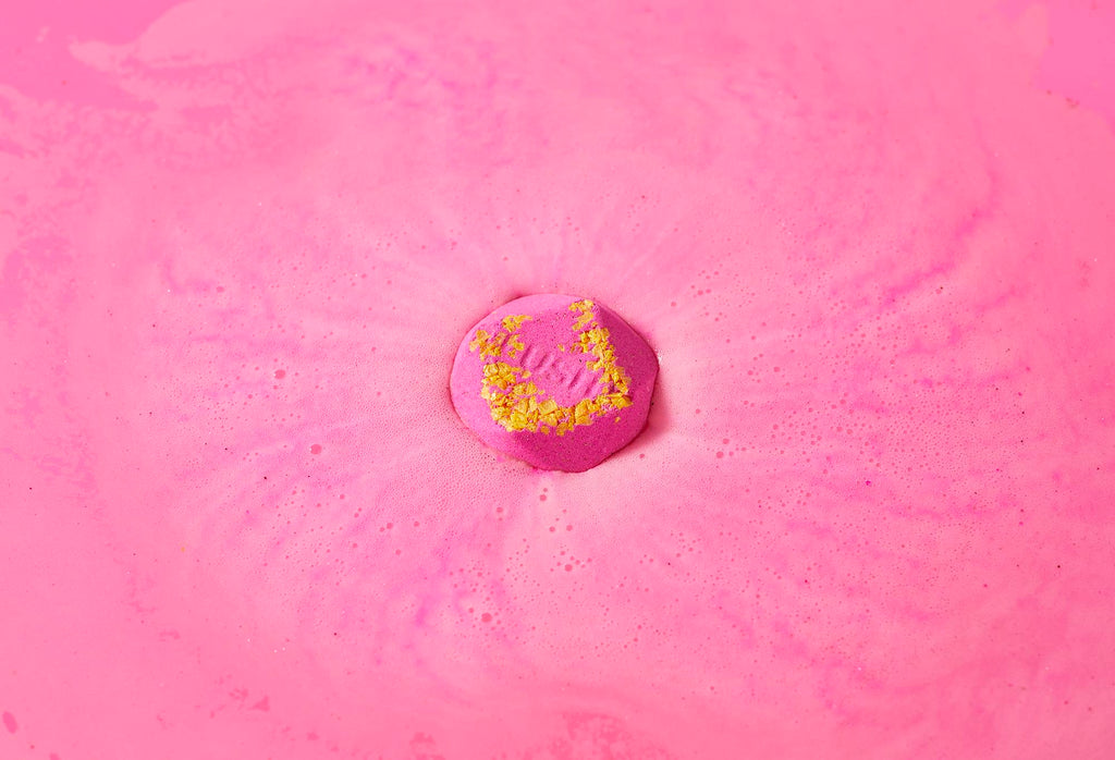 San San bath bomb sits on a sea of hot pink swirling foam with small pressed yellow-coloured salt and "Lush" embossed on top.