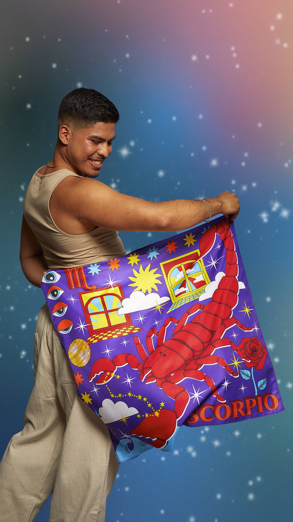 The image shows the model wearing a beige tank top and linen trousers. They are facing away on a pastel, sparkling background and holding the Scorpio knot wrap behind their back showing the full design.