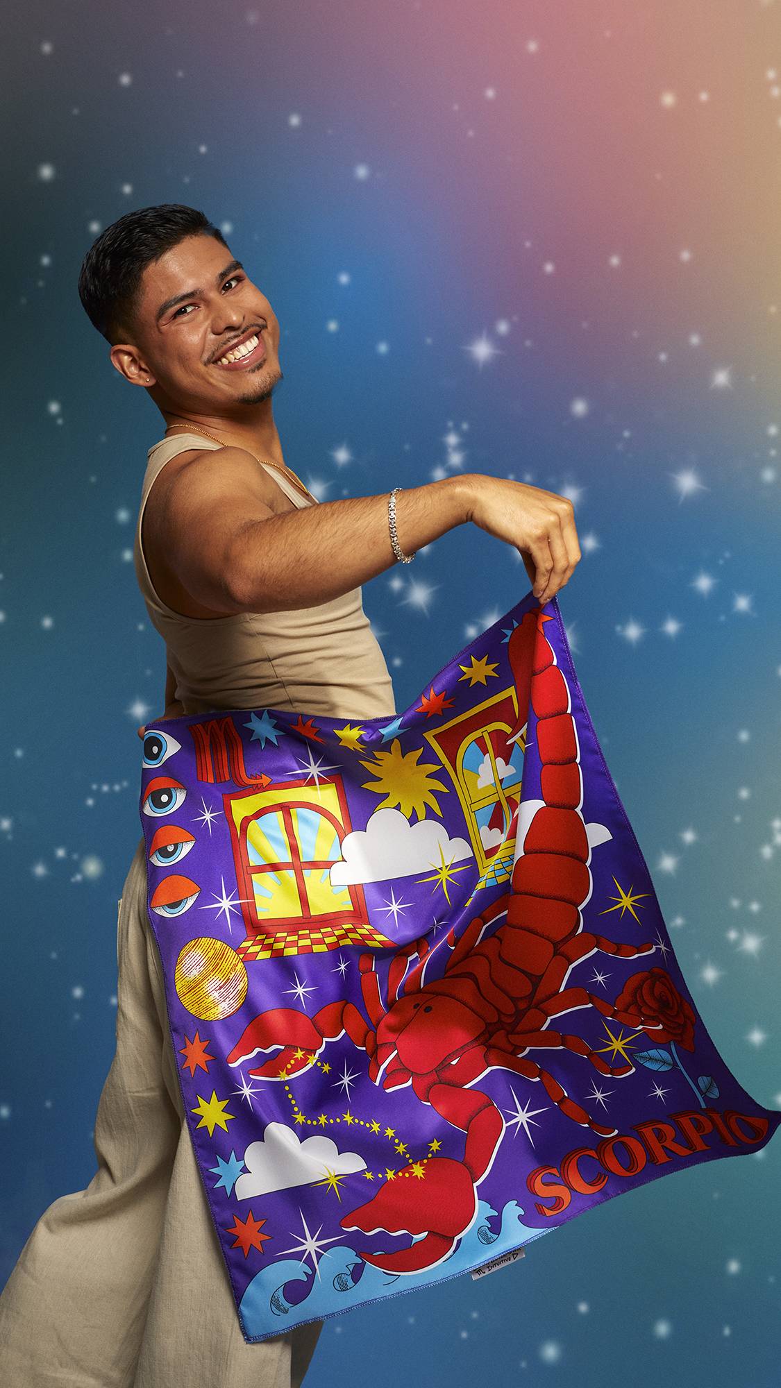 The image shows the model wearing a beige tank top and linen trousers. They are smiling on a pastel, sparkling background and holding the Scorpio knot wrap behind their back showing the full design.