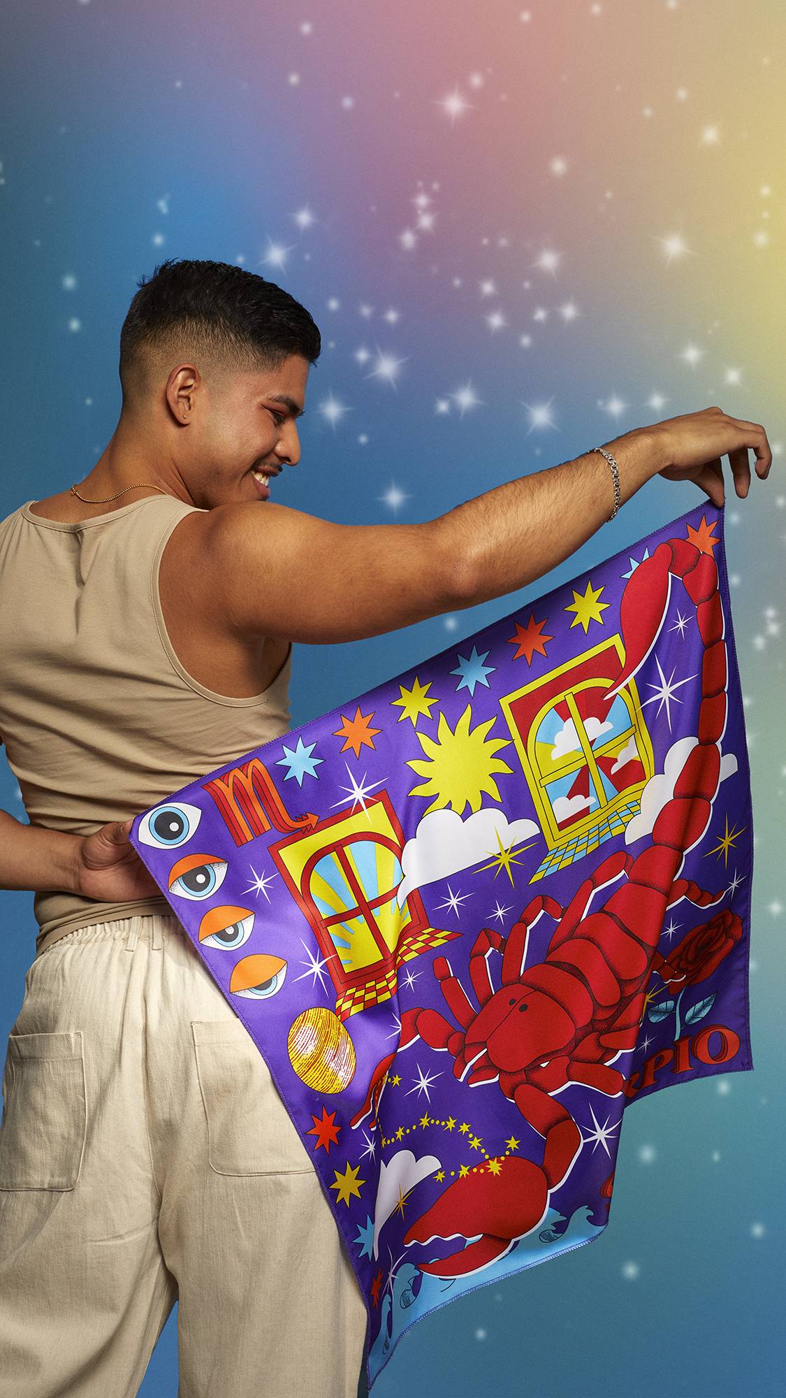 The image shows the model wearing a beige tank top and linen trousers. They are smiling on a pastel, sparkling background and holding the Scorpio knot wrap behind their back showing the full design.