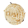 Soak and Float. A top-down shot of a circular, textured Lush solid shampoo bar. The cream-coloured bar features visible flecks of dried botanicals and has the word "LUSH" subtly stamped into its flaky surface.