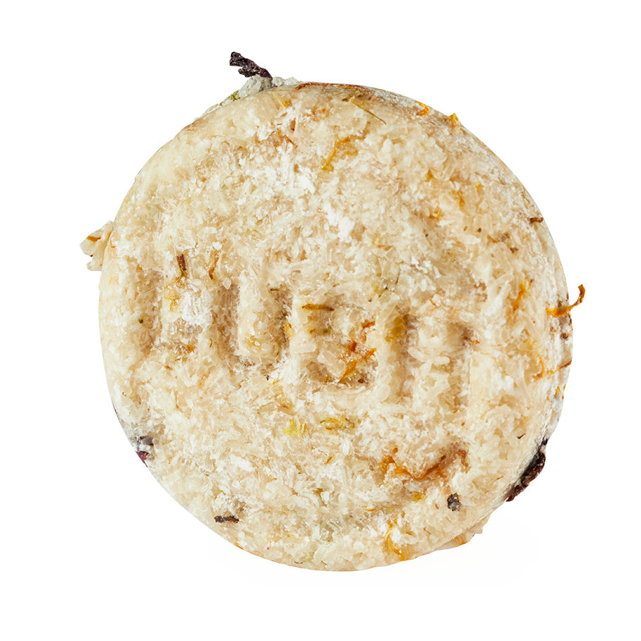 Soak and Float. A top-down shot of a circular, textured Lush solid shampoo bar. The cream-coloured bar features visible flecks of dried botanicals and has the word "LUSH" subtly stamped into its flaky surface.
