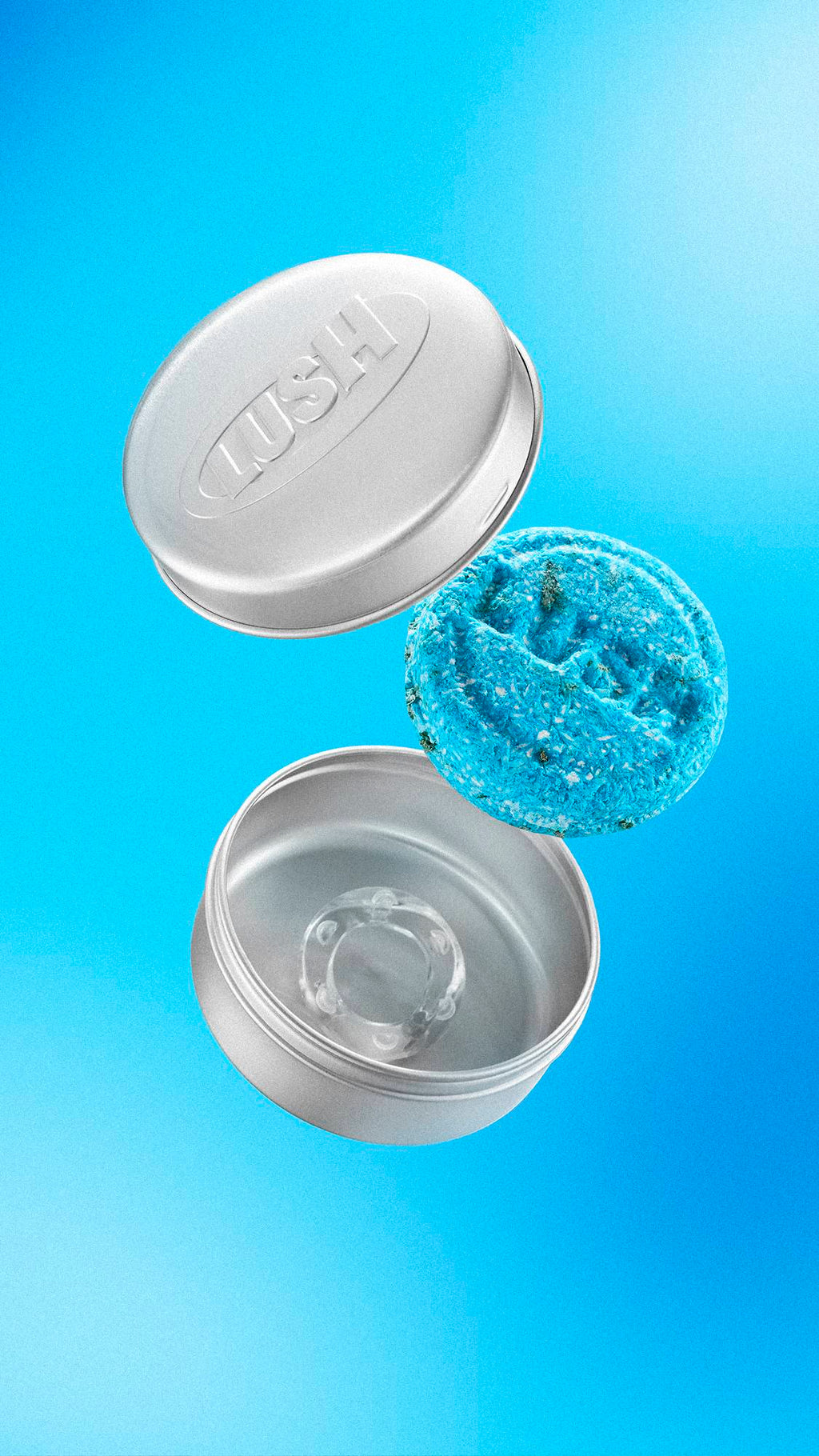 A textured, bright blue "Seanik" solid shampoo bar floats in mid-air between the lid and base of an open, silver aluminium Lush tin. The lid features an embossed 'LUSH' logo. Set against a light-to-mid blue gradient background.