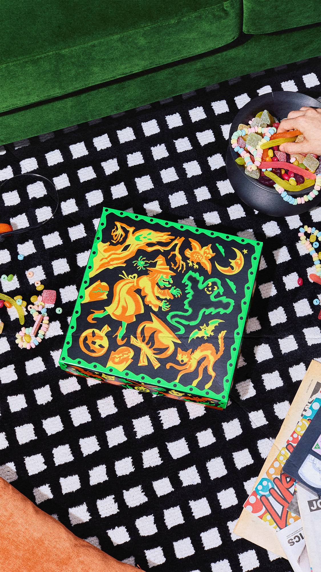 A black and green witch-themed gift box sits on a black and white chequered rug. A cauldron filled with sweets, loose candy necklaces, and a hand reaching for a treat are visible nearby, with a green couch in the background.