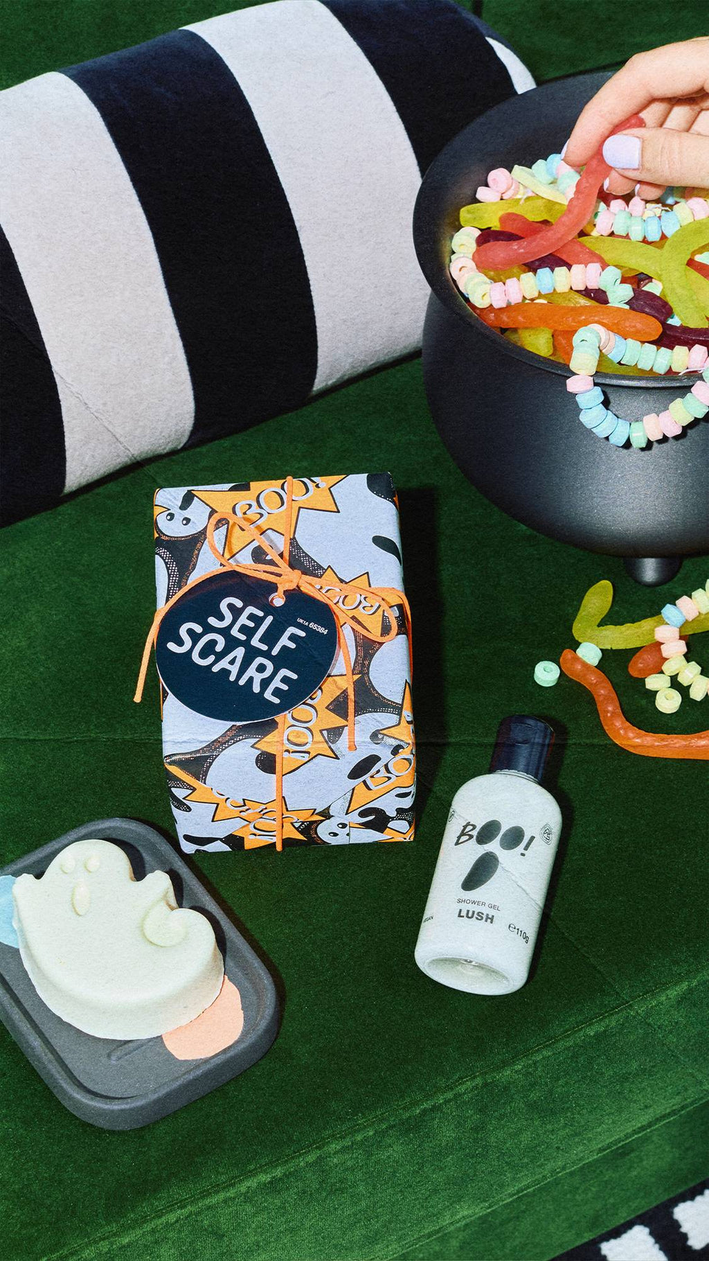 A Lush 'Self Scare' wrapped gift box, 'BOO!' shower gel, and a ghost-shaped soap on a green velvet surface. A black cauldron filled with colourful sweets and a striped cushion sit nearby as a hand reaches for a sweet.
