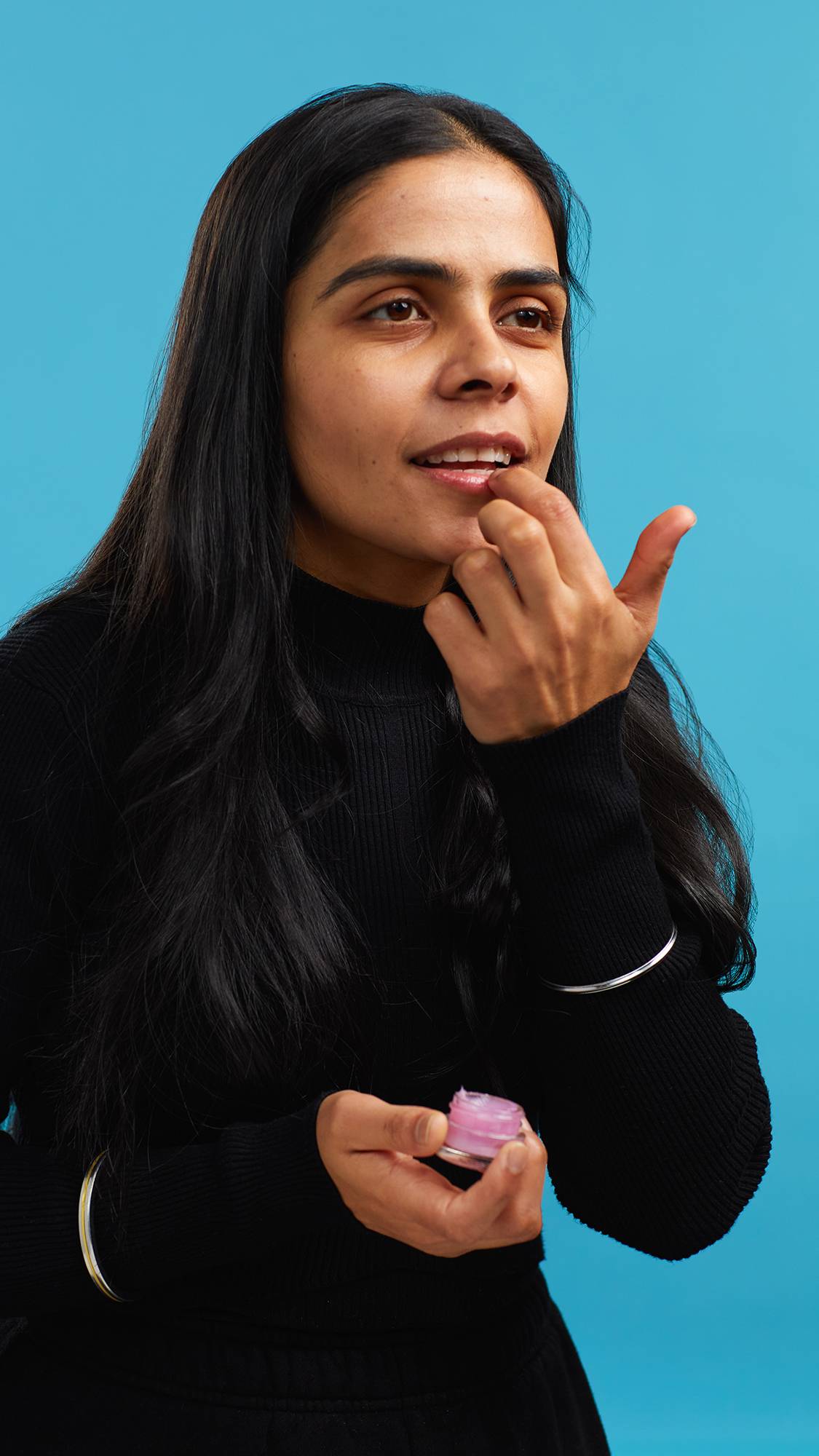 A close-up shot of the model on a sky-blue background wearing a black turtleneck. They are holding a small, glass pot of the pink Sex Bomb lip gloss as they apply it to their lips.