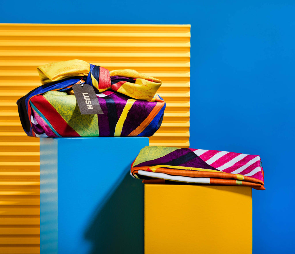 The Knot Wrap is decoratively wrapped with a Lush gift tag, alongside one folded, in front of a blue and yellow background.