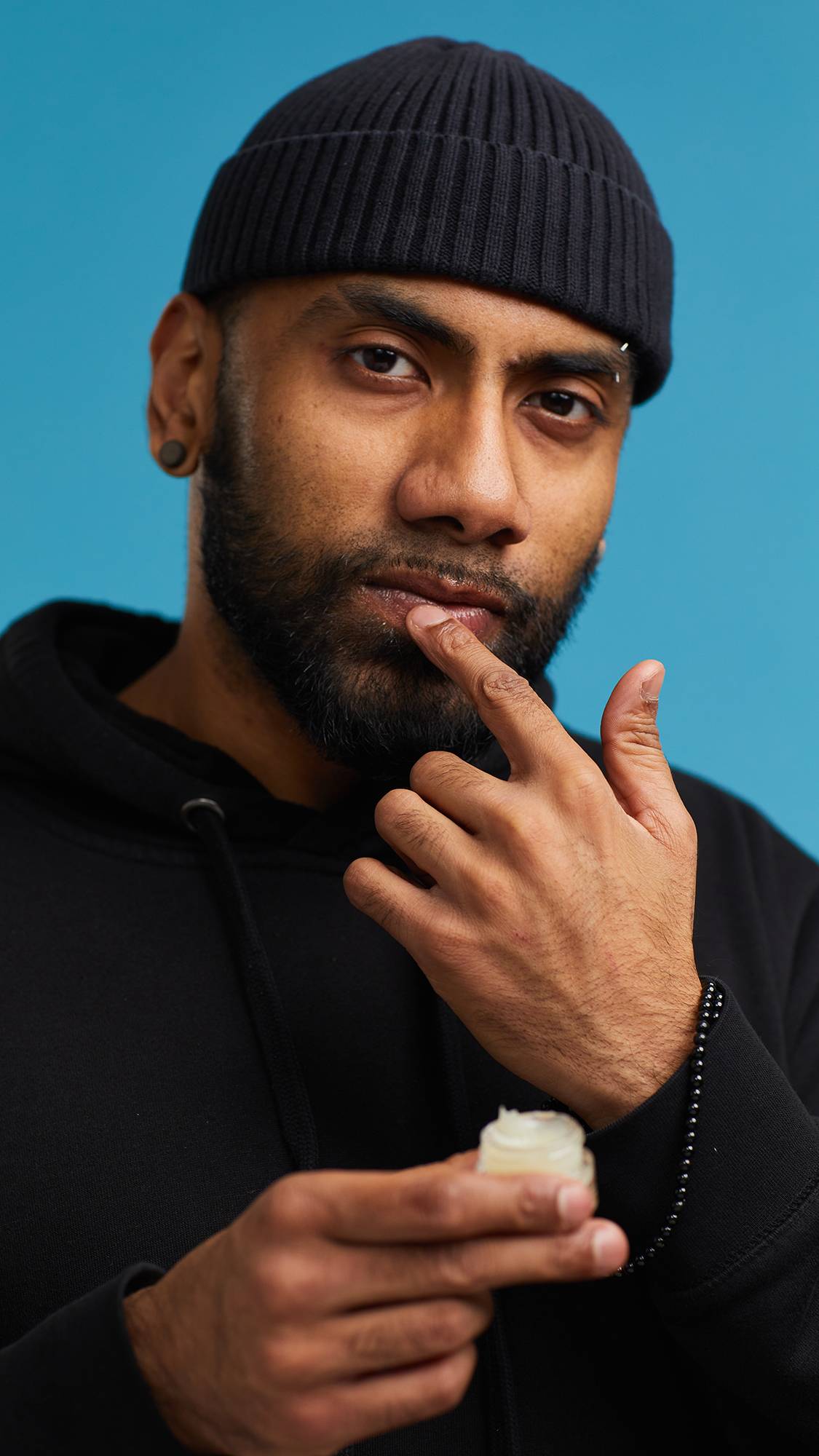 A close-up shot of the model on a sky-blue background wearing a black hoodie and hat. They are holding a small, glass pot of the Shimmy Shimmy lip gloss as they apply it to their lips.