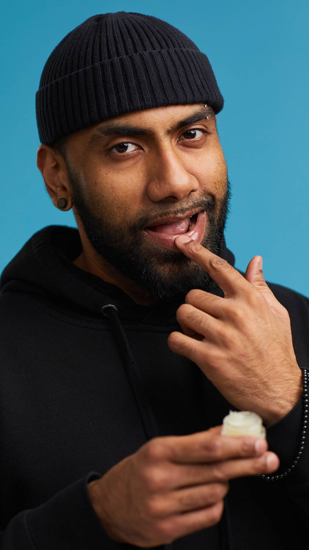 A close-up shot of the model on a sky-blue background wearing a black hoodie and hat. They are holding a small, glass pot of the Shimmy Shimmy lip gloss as they apply it to their lips.