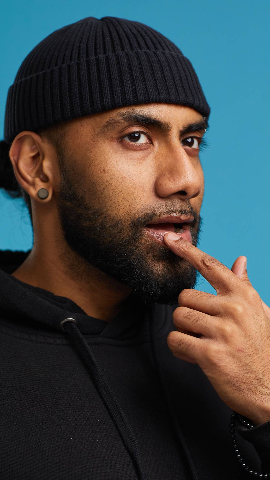 A close-up shot of the model on a sky-blue background wearing a black hoodie and hat. They are applying the Shimmy Shimmy lip gloss with the tip of their index finger.