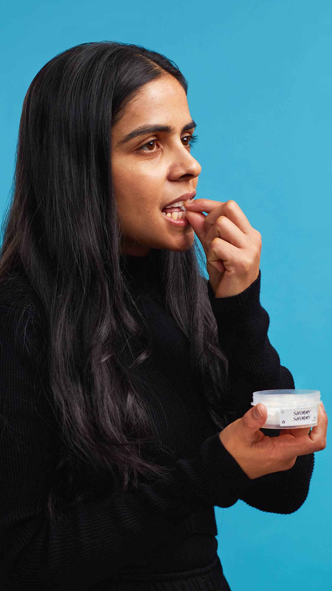 The image shows the model wearing a black turtleneck on a sky-blue background. They are holding a small, open clear, recycled plastic tub of Shimmy Shimmy mouthwash tabs and gently placing one into their mouth.