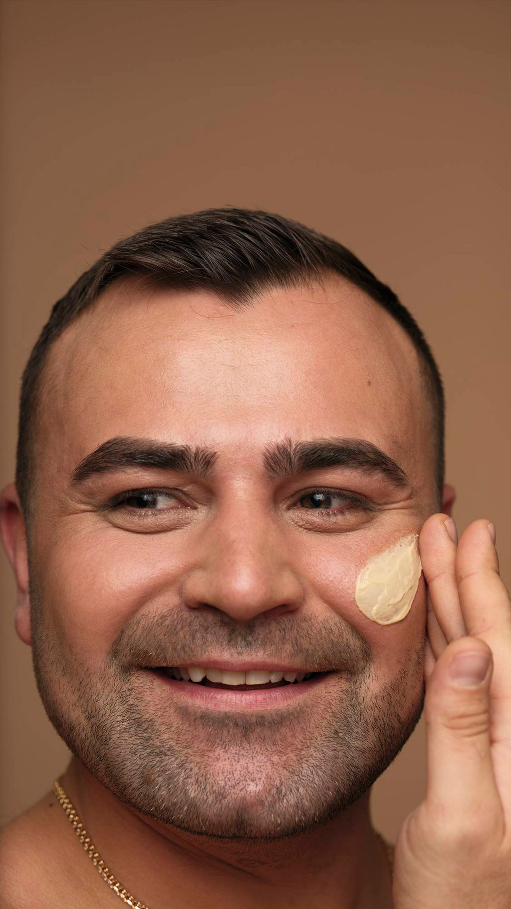 A close-up photograph of a smiling person with short dark hair and facial hair applying Skin Drink moisturiser to their cheek with their fingers against a neutral background.