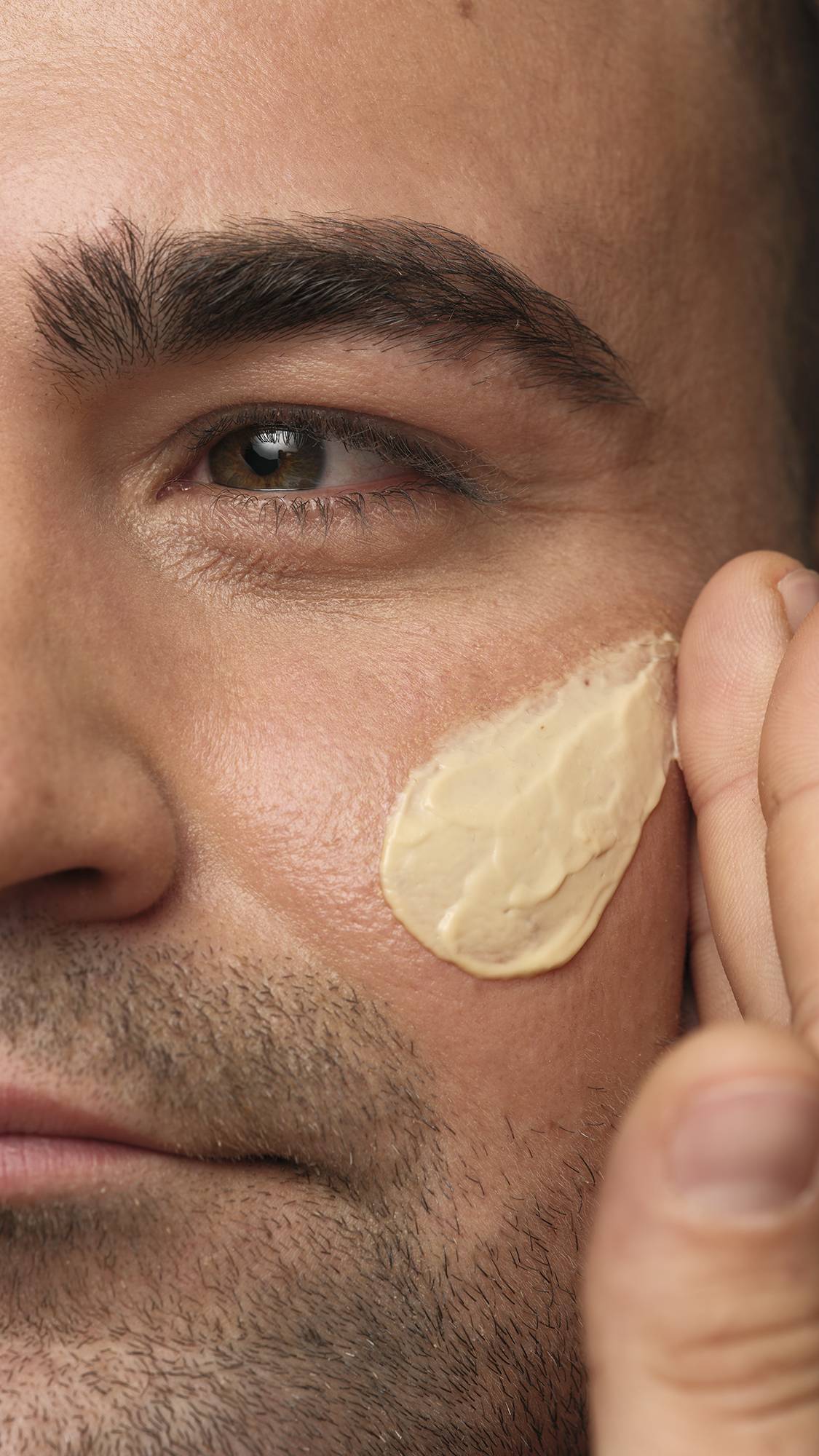 A close-up portrait of a smiling person with short dark hair and facial stubble looking directly at the camera, with dabs of Skin Drink moisturiser applied to both cheeks against a neutral background.