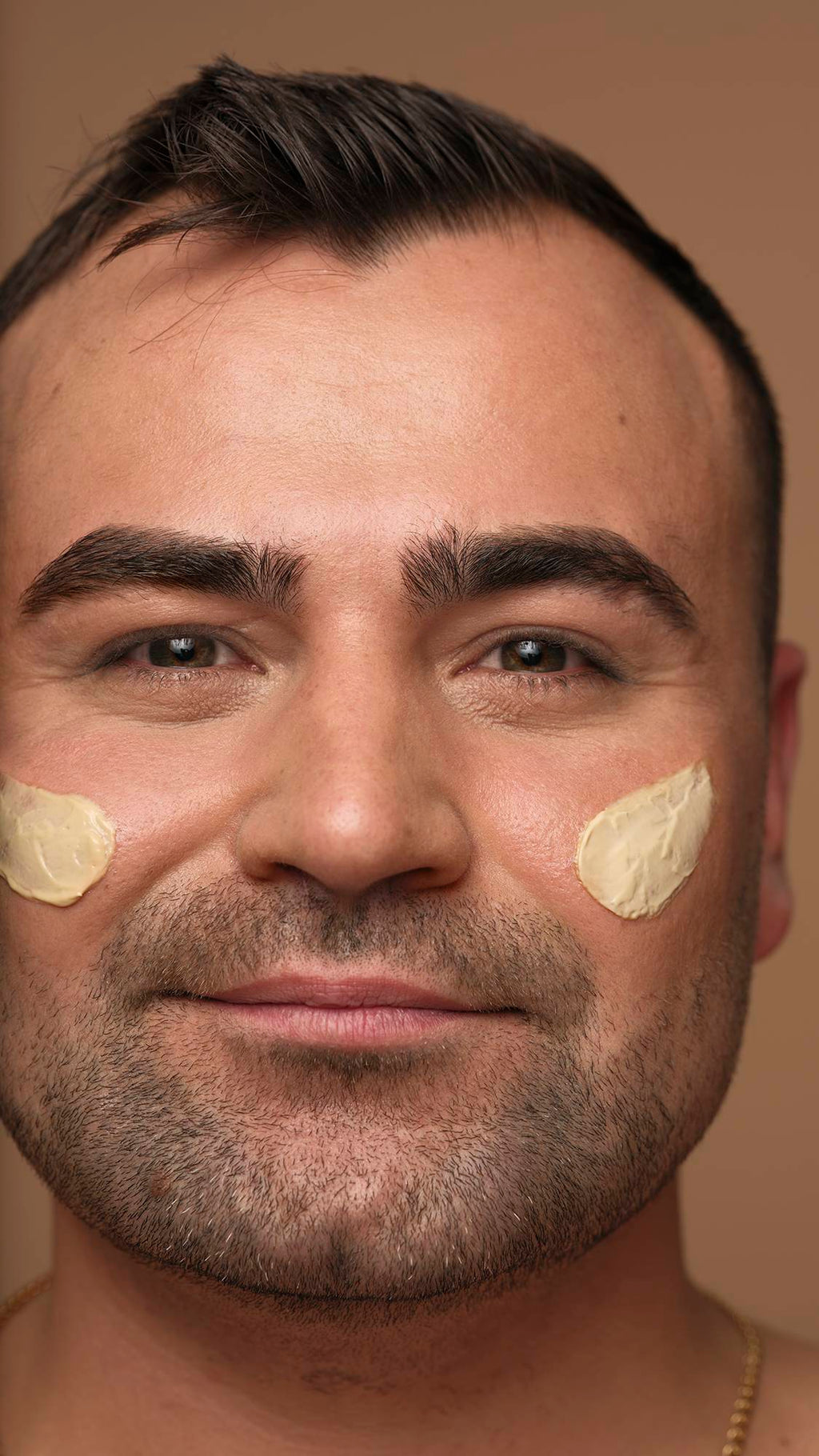 A close-up portrait of a smiling person with short dark hair and facial hair looking directly at the camera, with dabs of Skin Drink moisturiser applied to both cheeks against a neutral background.