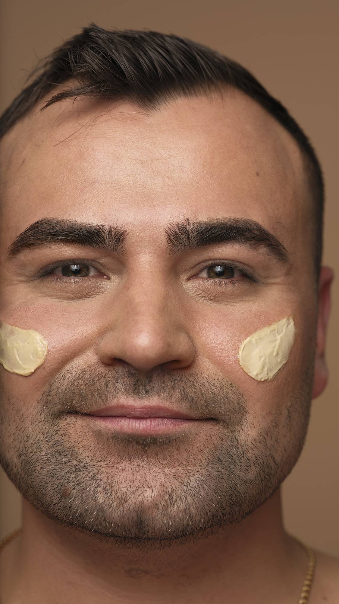 A close-up portrait of a smiling person with short dark hair and facial hair looking directly at the camera, with dabs of Skin Drink moisturiser applied to both cheeks against a neutral background.