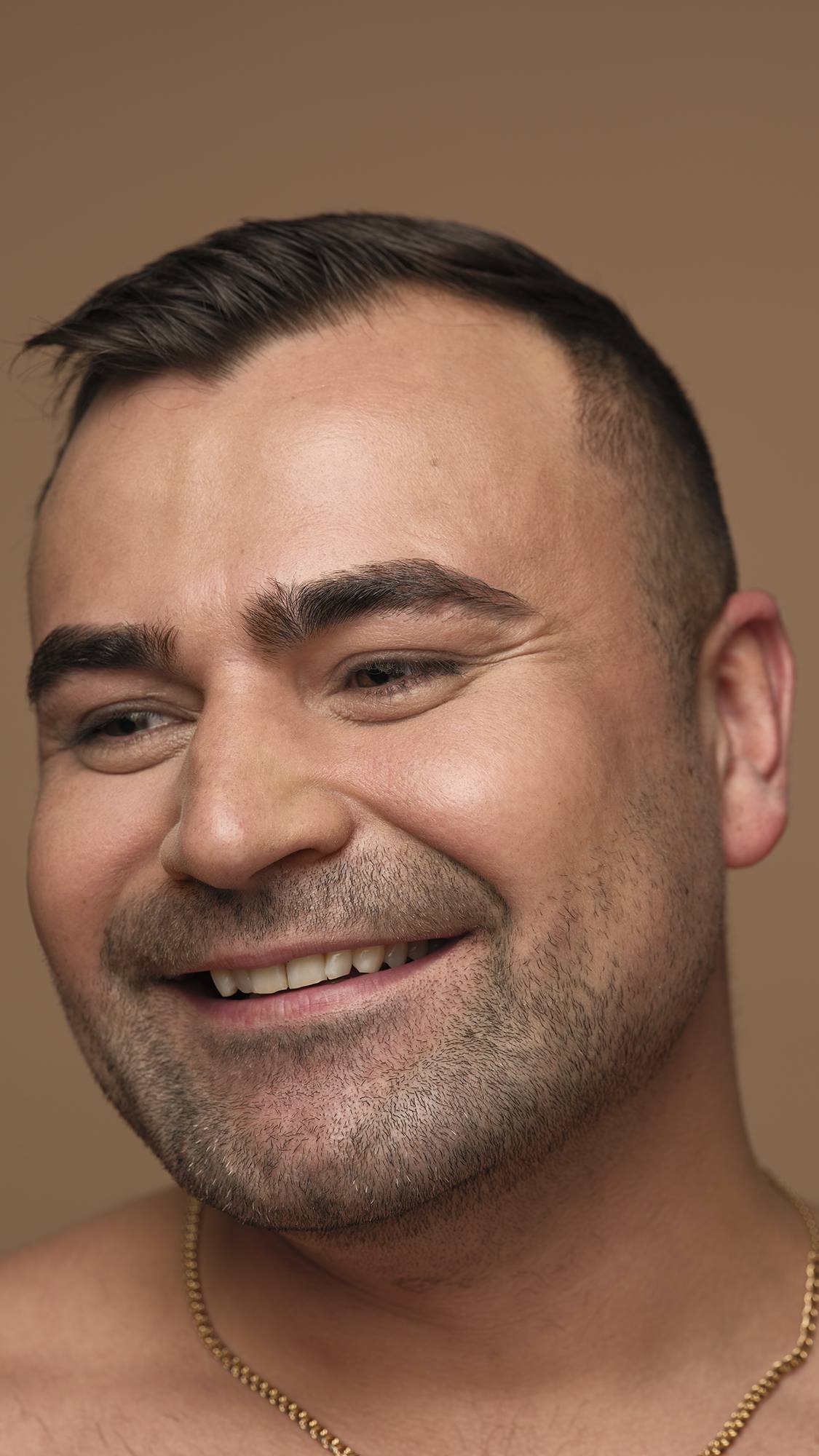 A close-up side profile of a smiling person with short dark hair and facial stubble, wearing a gold chain and looking off-camera against a neutral background.