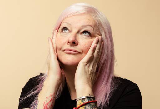 A person with light pink hair looks up nonchalantly, as they push their cheeks upwards with their hands covered in moisturiser.