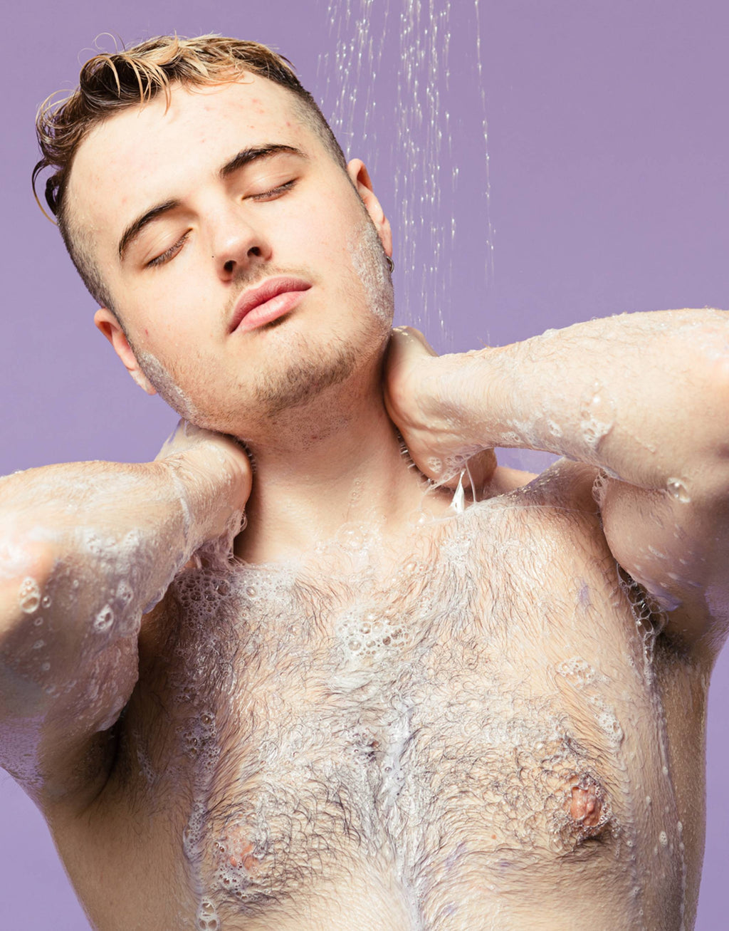 The image shows a model on a pastel, lavender-purple background while under running shower water. They have applied the lavender, Sleepy shower gel to their body and are lathering up soapy suds over their neck and chest.
