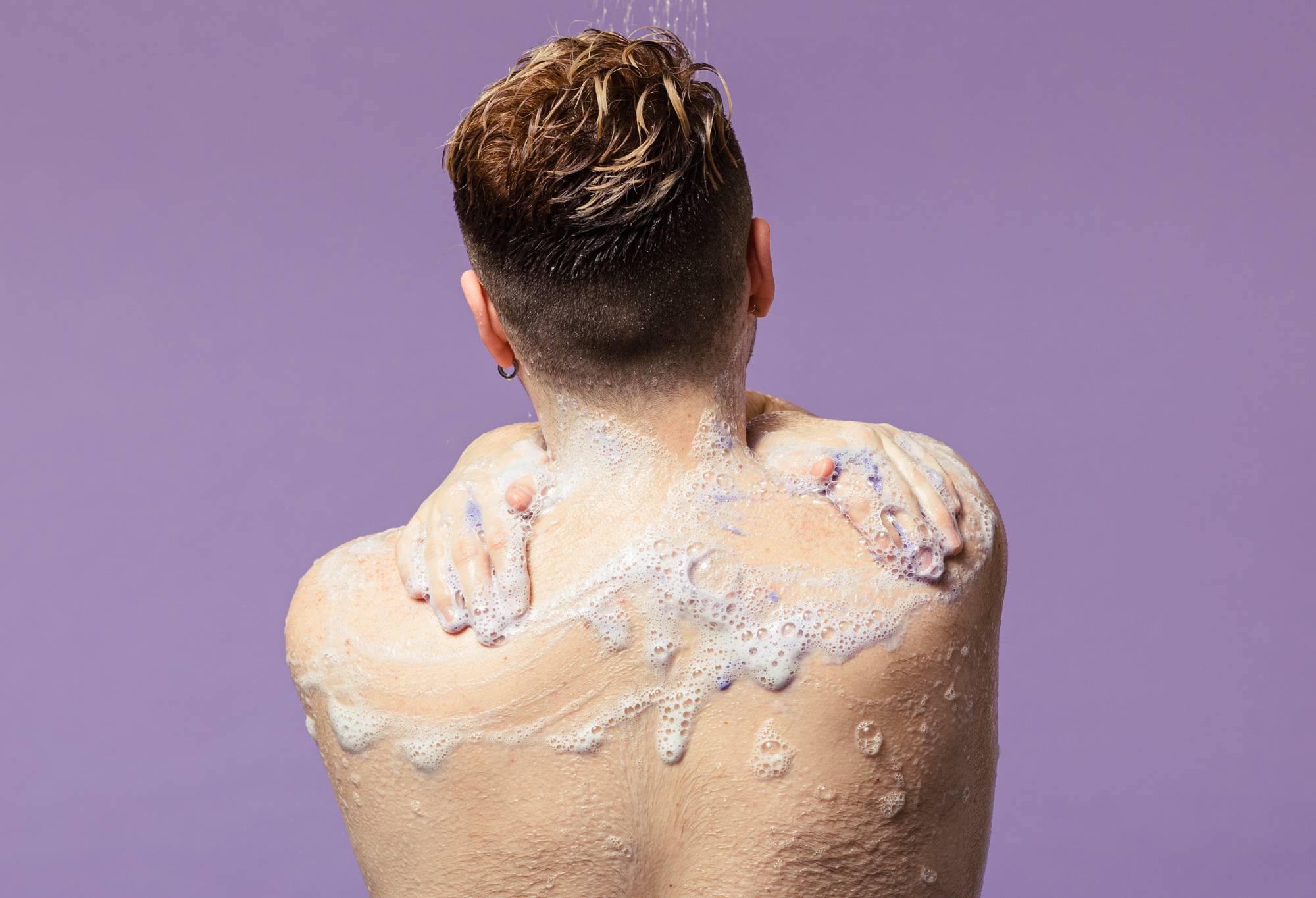 Seen from behind, a person with short, damp hair washes in the shower, lathering Lush "Sleepy" shower gel over their shoulders and upper back. A subtle hint of purple pigment peeks through the white foam against a solid pastel lilac background.