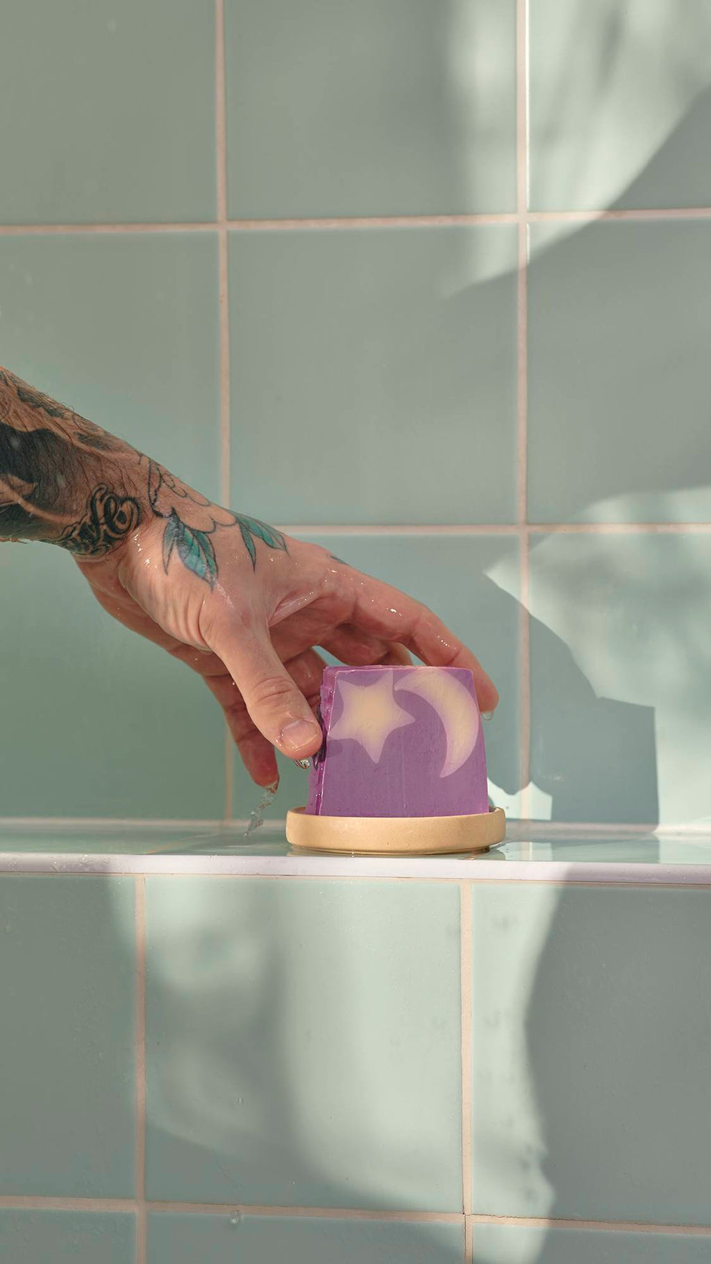 A tattooed hand reaches for a purple Sleepy soap bar with a star and moon design. The soap sits in a dish on a light green tiled shower ledge.