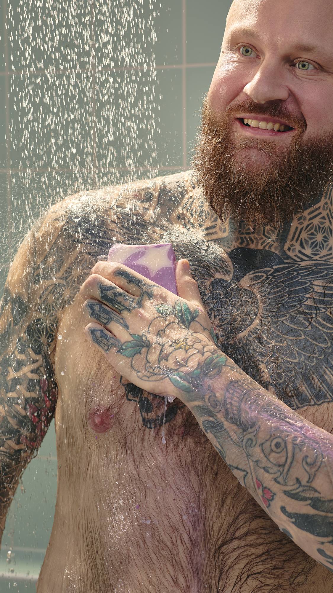 A smiling man with chest and arm tattoos uses a purple Sleepy soap bar under a shower spray. The soap features a white star design, and the background shows light green tiles.