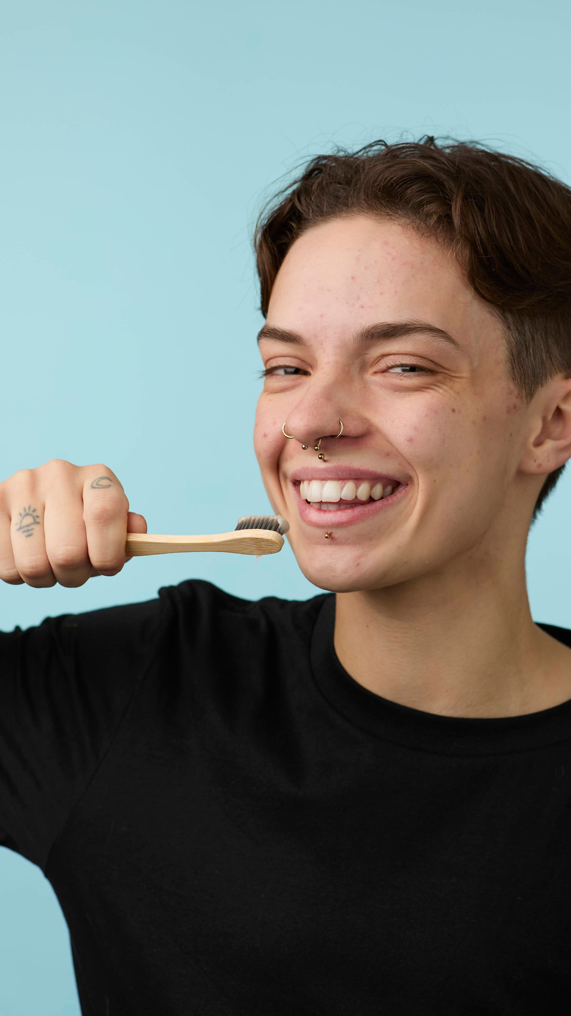 The image shows the model in a back t-shirt on a pale blue background. They are smiling widely as they hold a bamboo toothbrush loaded with the LUSH Sleepy toothpaste to their face.