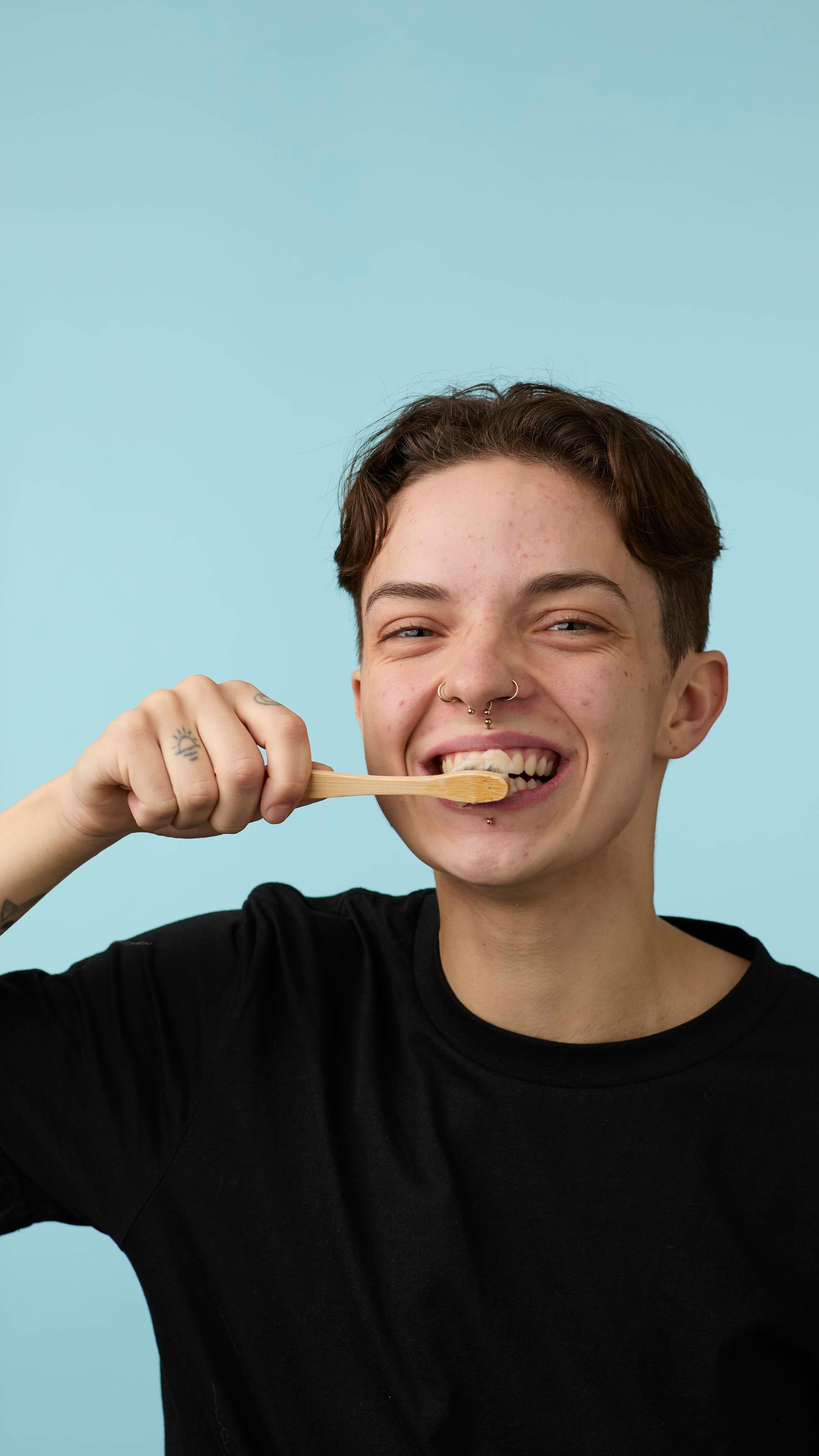 The image shows the model in a back t-shirt on a pale blue background. They are smiling widely as they use a bamboo toothbrush loaded with the LUSH Sleepy toothpaste and begin to brush.