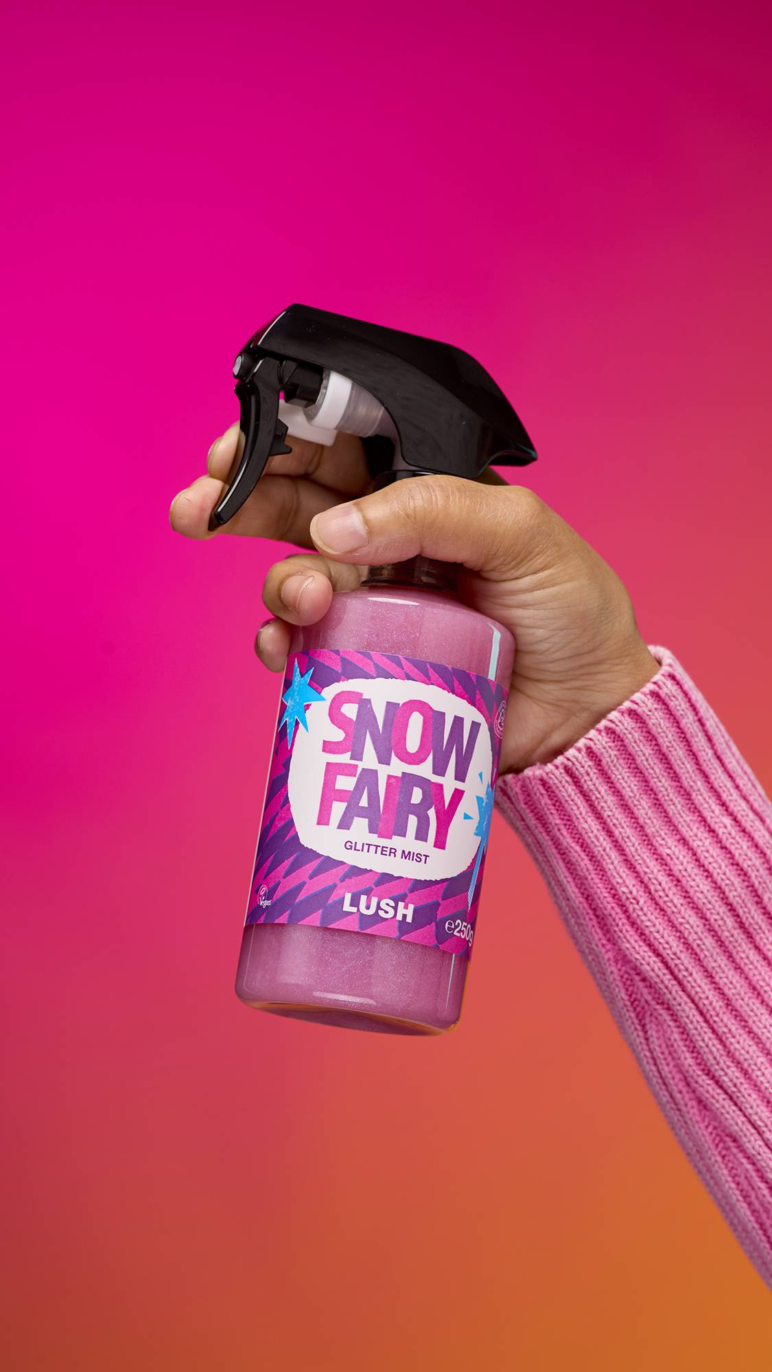 An arm reaching from the right-hand side. The hand holds a transparent, cylindrical plastic spray bottle with pink, shimmering liquid and a black top. A colourful label reads "Snow Fairy Glitter Mist" on a pink-to-orange gradient background.