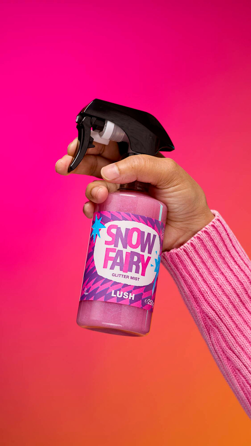 An arm reaching from the right-hand side. The hand holds a transparent, cylindrical plastic spray bottle with pink, shimmering liquid and a black top. A colourful label reads "Snow Fairy Glitter Mist" on a pink-to-orange gradient background.
