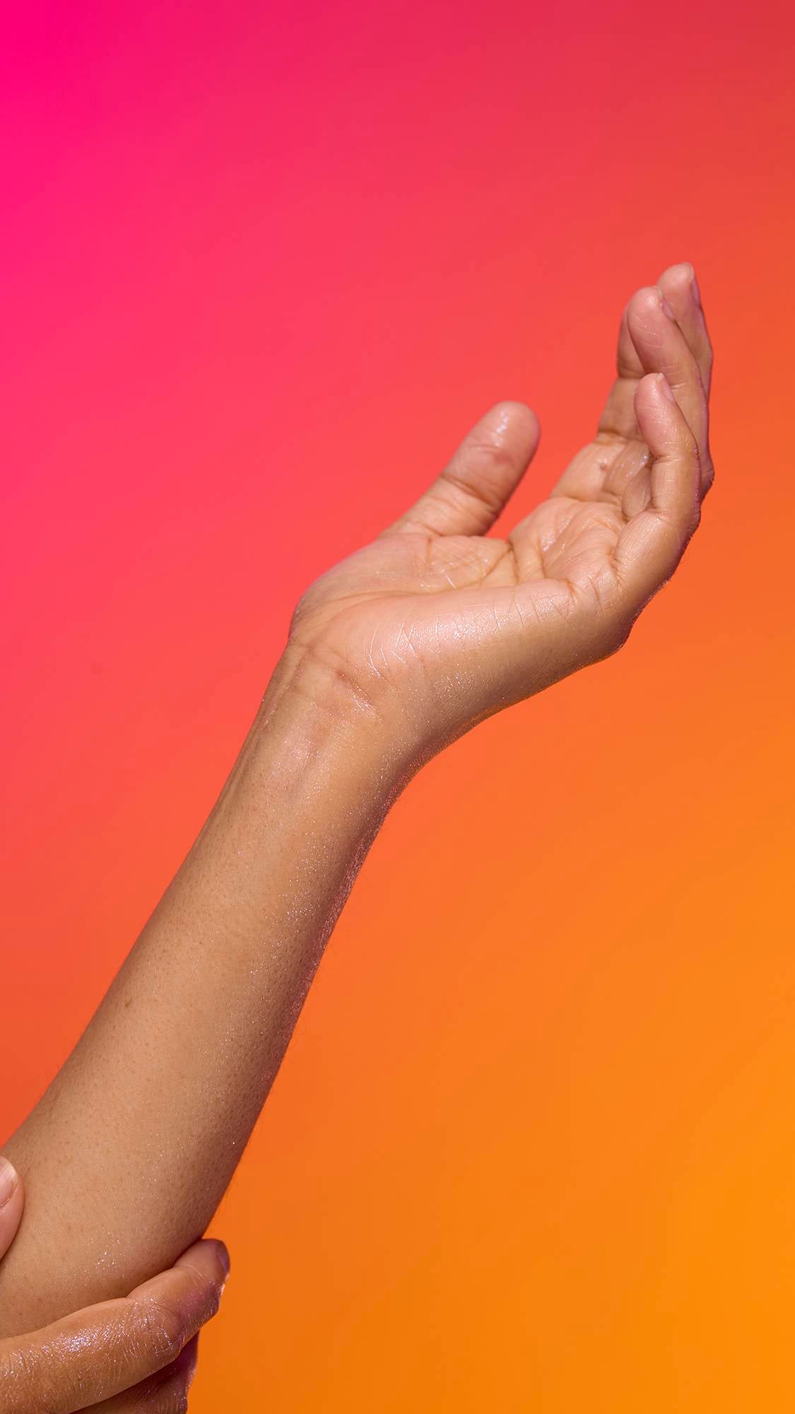The image shows a model's forearm reaching in from the left-hand side as the skin appears moisturised with a subtle, delicate shimmer over this skin after having used the Snow Fairy Glitter Mist spray product on a pink-to-orange gradient background.