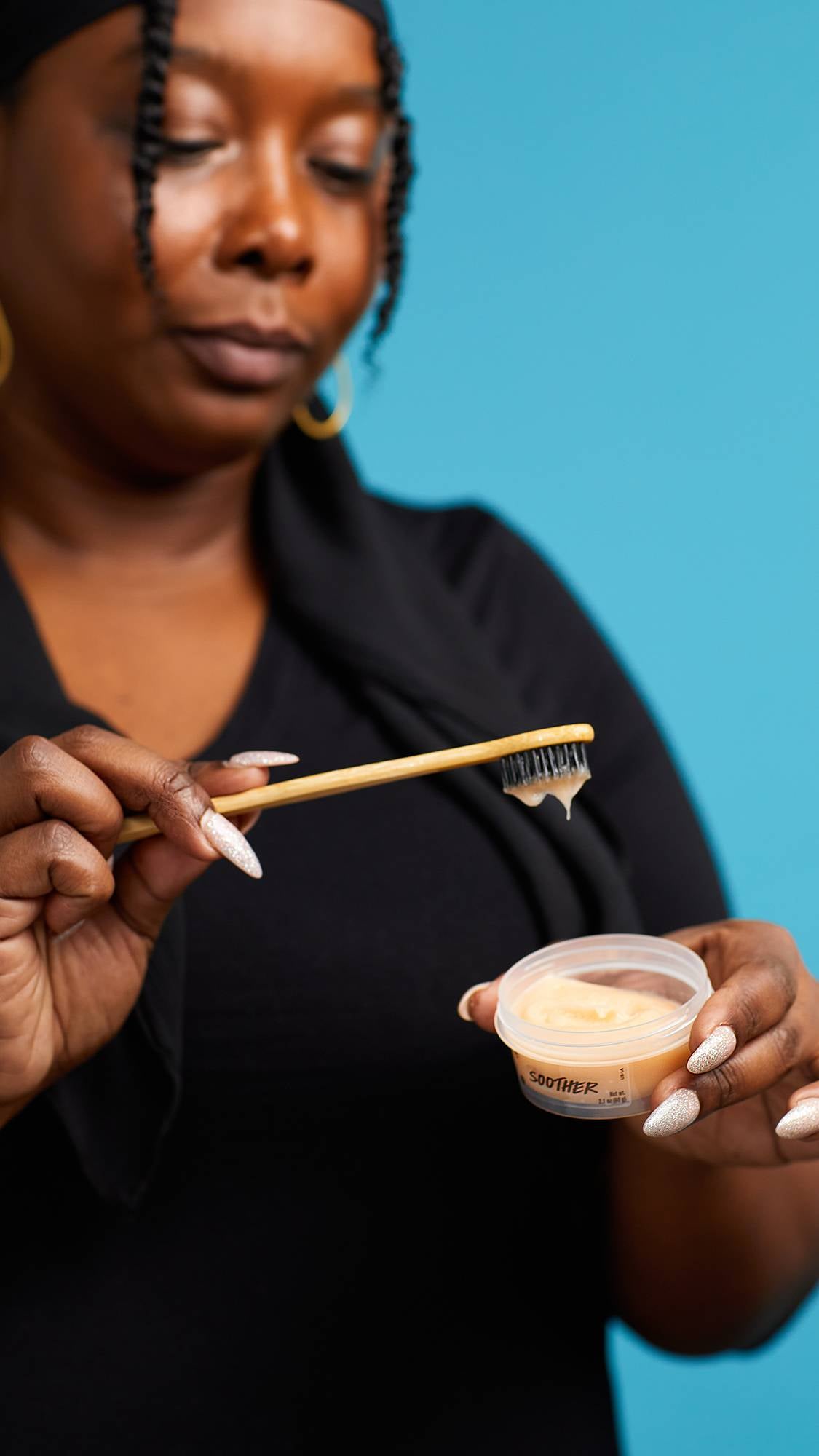 The image shows the model in a black top on a sky-blue background. They are holding an open, clear, recycled plastic pot of Soother toothpaste as they dip the head of their toothbrush into the pot with the other hand.