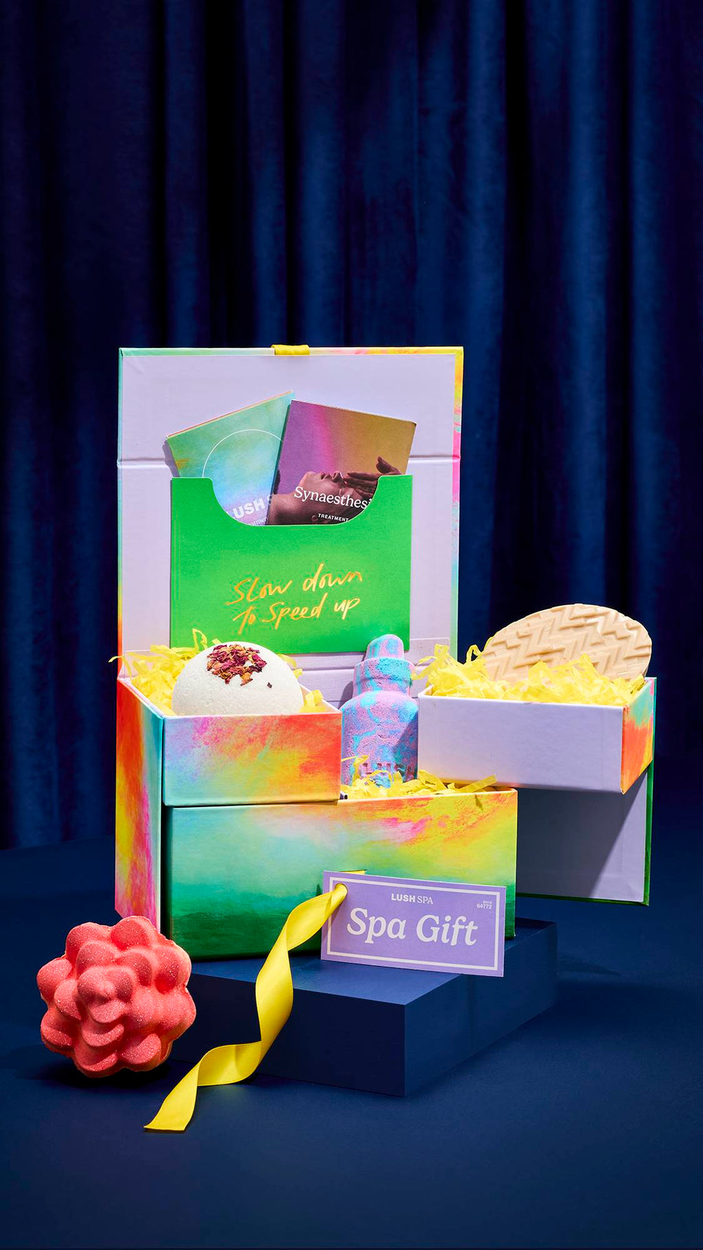 An open LUSH SPA gift box with multiple tiers reveals a white dome-shaped solid body scrub, a marbled blue and pink bottle-shaped bath bomb, and a massage bar. A pink flower-shaped bath bomb sits on the table beside the colourful box.