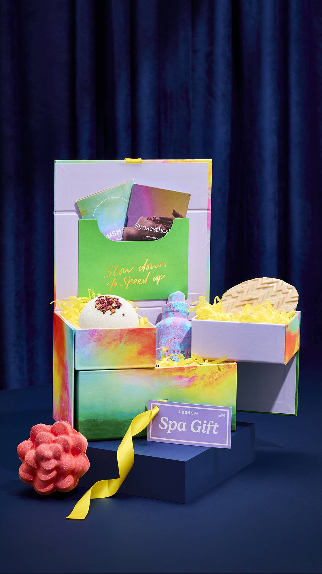 An open LUSH SPA gift box with multiple tiers reveals a white dome-shaped solid body scrub, a marbled blue and pink bottle-shaped bath bomb, and a massage bar. A pink flower-shaped bath bomb sits on the table beside the colourful box.