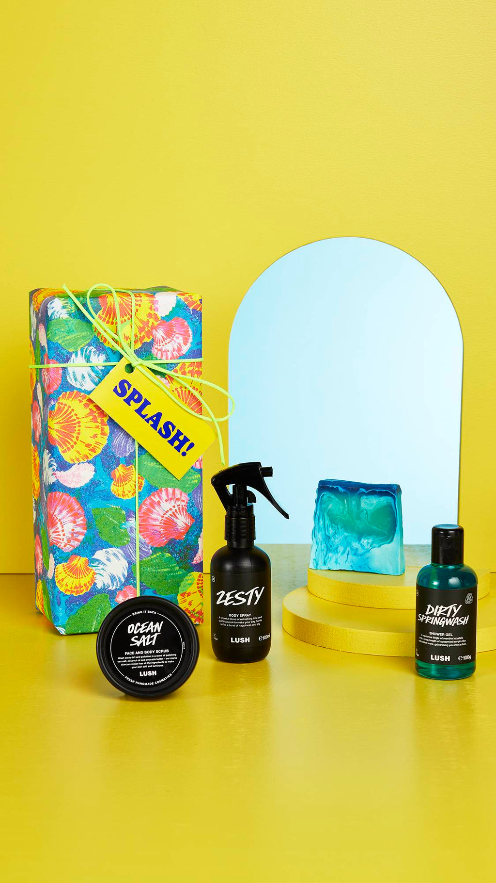 The shell-patterned gift box and the four LUSH products sit on a bright yellow floor and background with an arched mirror in the background.