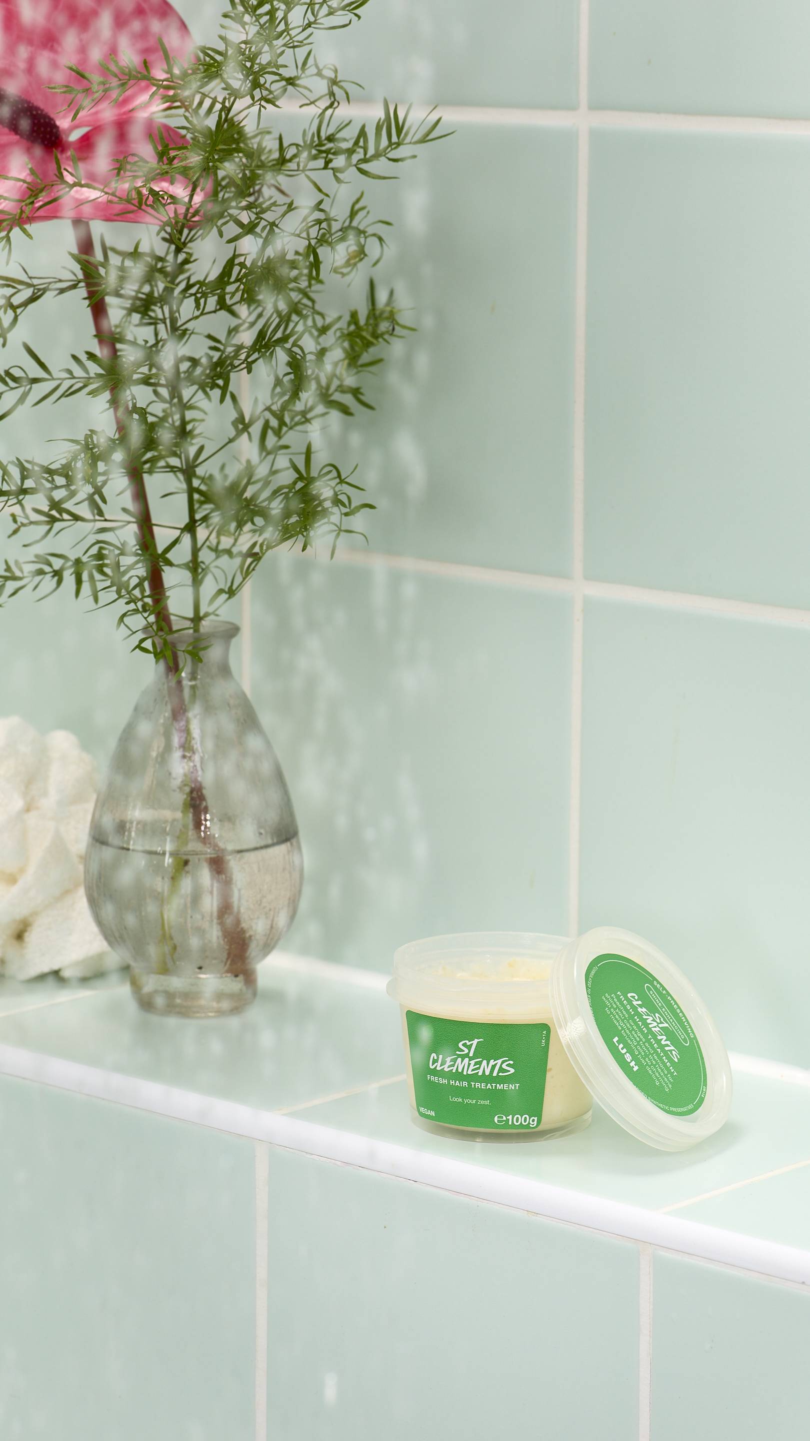 An open pot of pale yellow "St Clements" fresh hair treatment by Lush sits on a light green tiled ledge. In the background, a glass vase holds a green plant and a pink flower, partially obscured by falling water droplets.