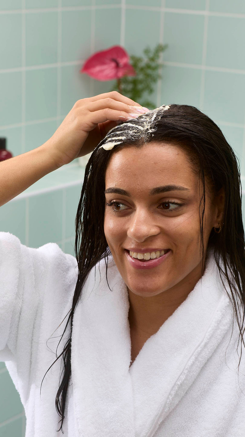 The image shows a model smiling while applying a pale cream-coloured "St Clements" hair treatment mask to their hair parting. They are wearing a white dressing gown, with a blurred pink flower and green bathroom tiles visible in the background.