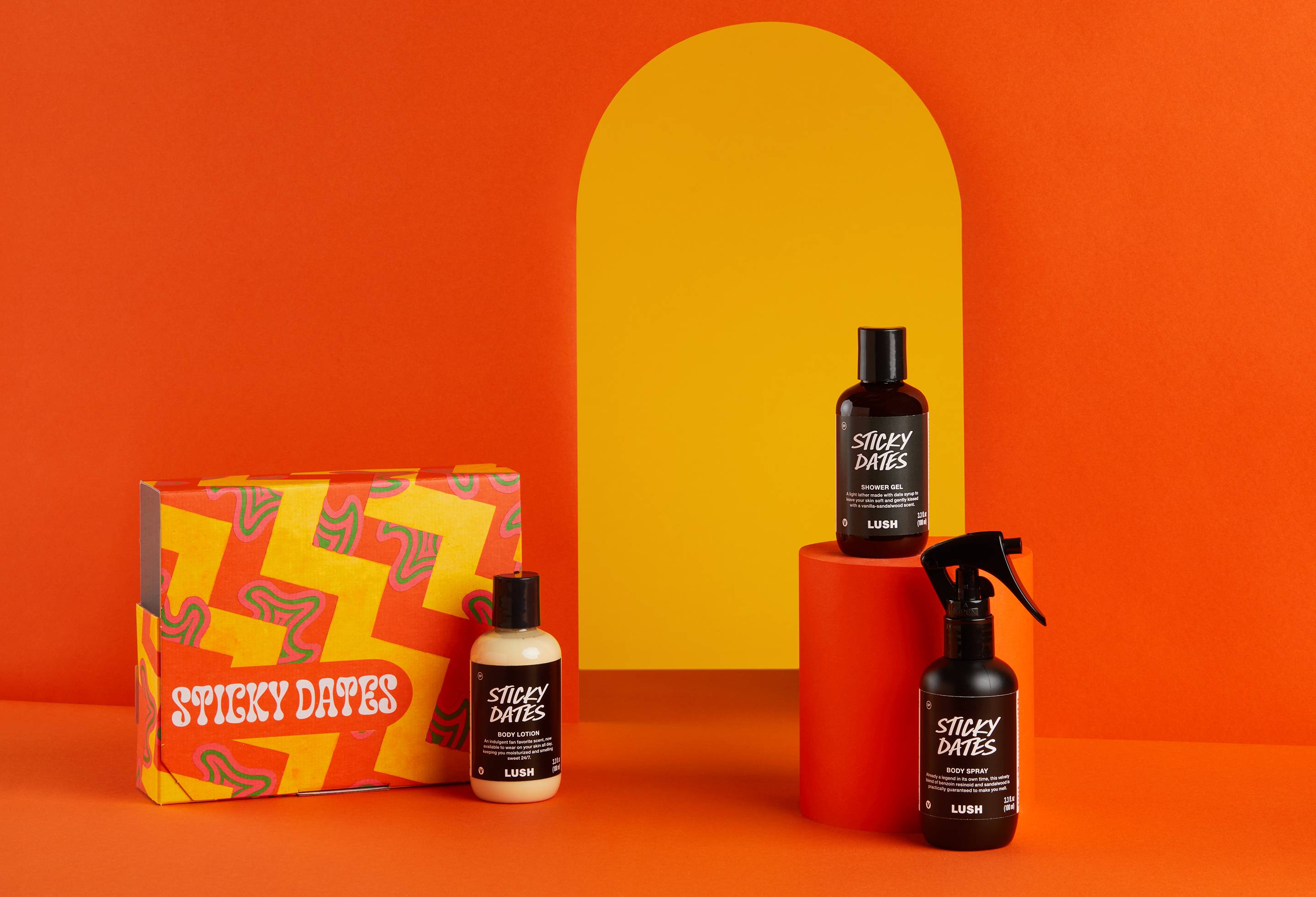 The image shows a bright orange background and flooring with a yellow arch detail/ The Sticky Dates gift box sits on the floor surrounded by the three included Sticky dates products. There is a body lotion, shower gel and body spray.