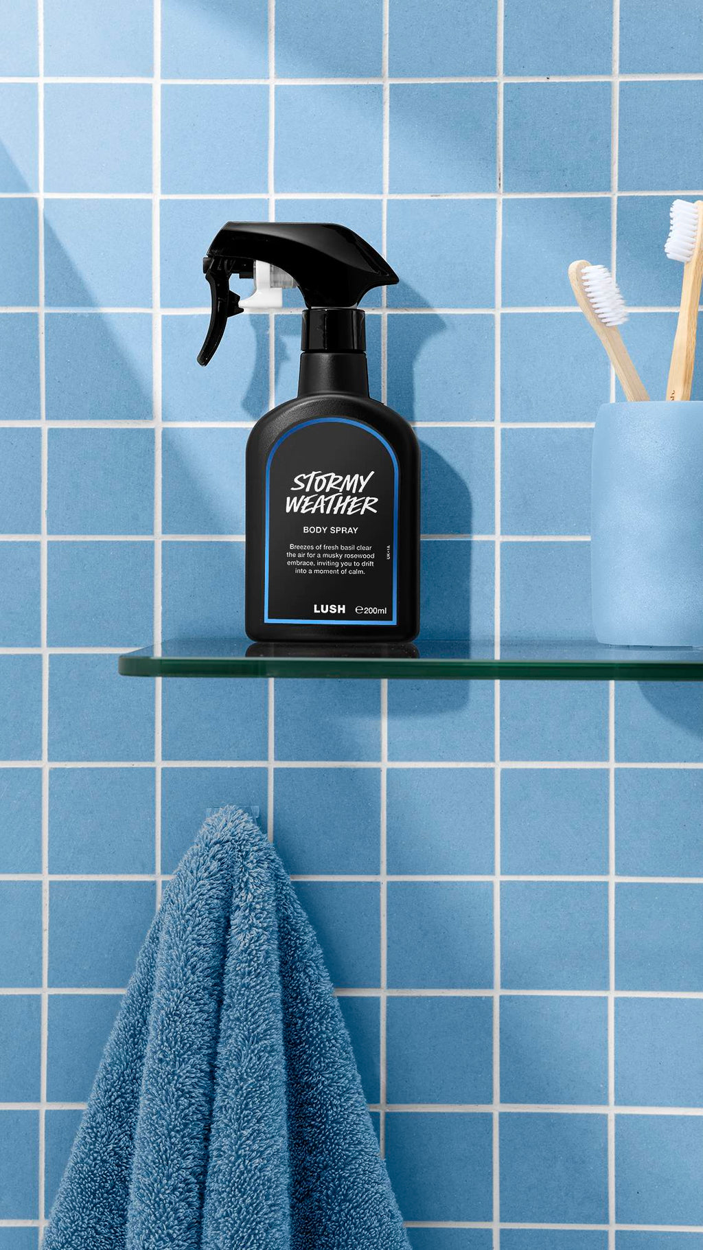 The image shows a bottle of LUSH 'Stormy Weather' body spray on a glass shelf. A blue towel hangs below, with a blue cup and toothbrushes nearby, against a bright, tiled, sky-blue bathroom wall.