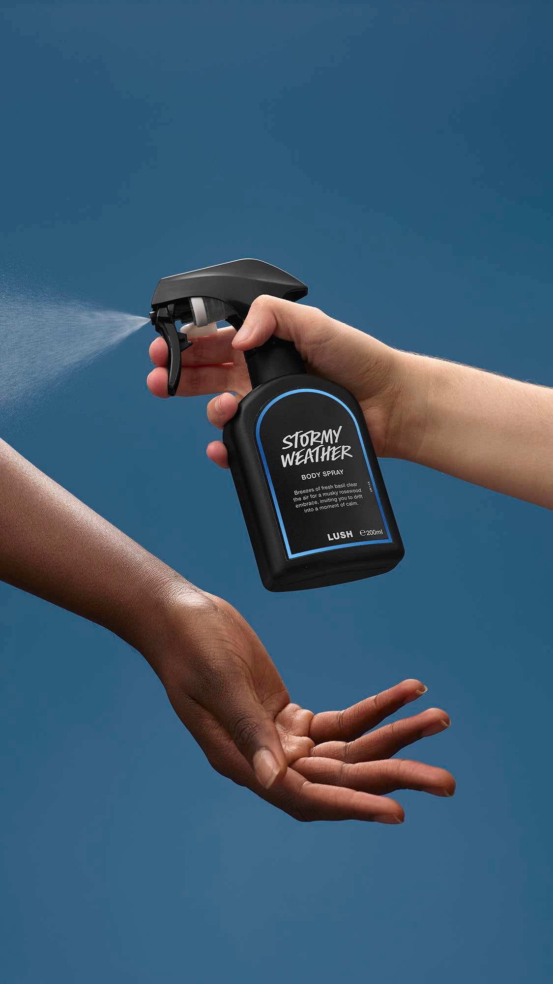 The image shows a light-toned hand spraying a bottle of LUSH 'Stormy Weather' body spray. A warm-toned hand is cupped below, catching the fine mist against a sea-blue background.