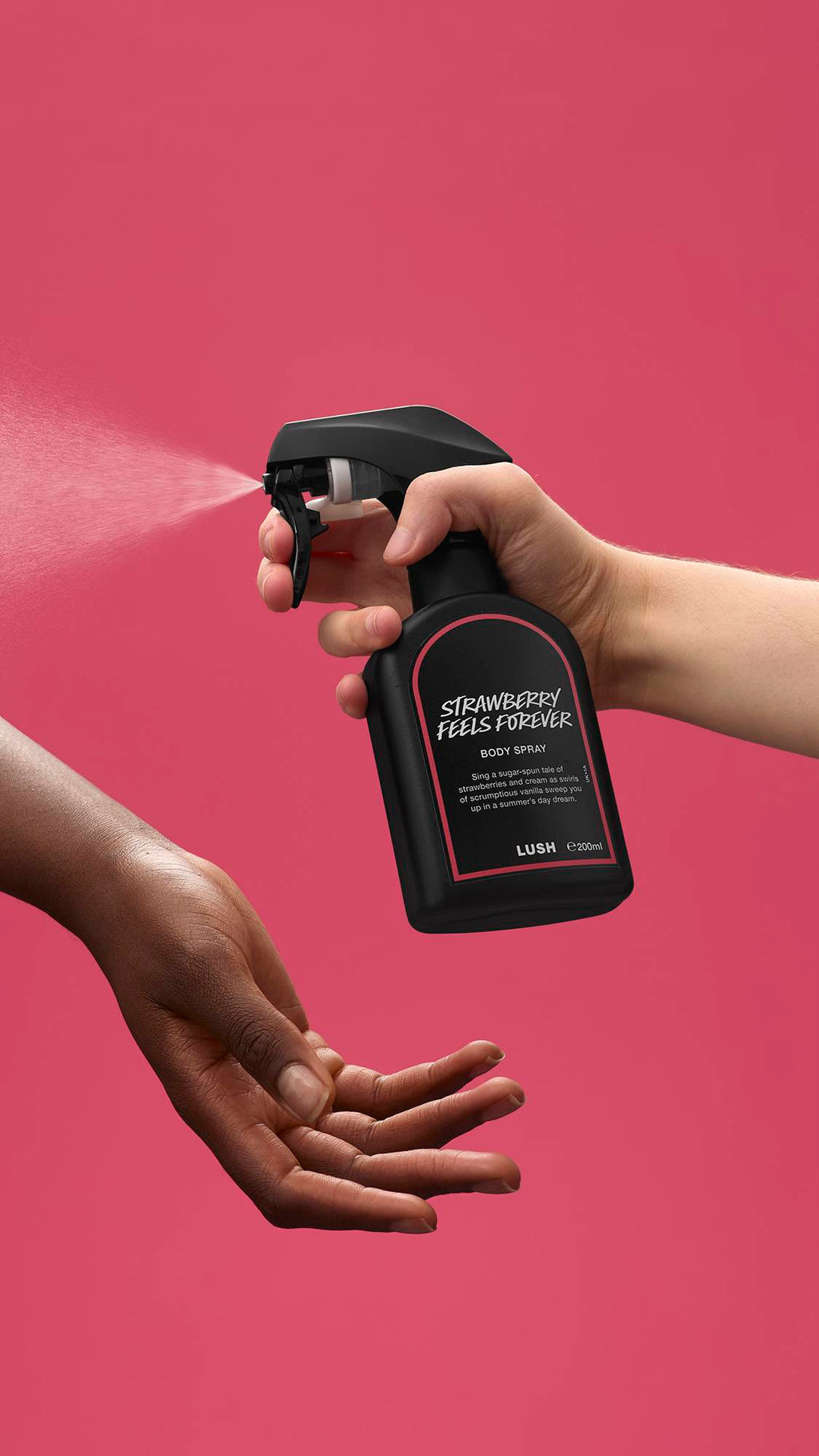 The image shows a light-toned hand spraying a bottle of LUSH 'Strawberry Feels Forever' body spray. A warm-toned hand is cupped below, catching the fine mist against a deep, rose-pink background.